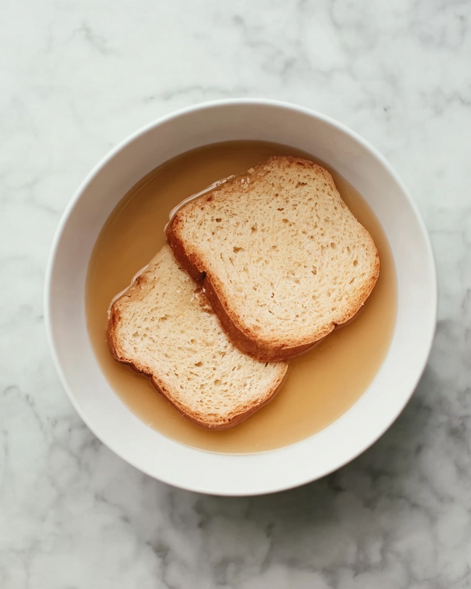 A white bowl filled with light brown liquid, inside which two slices of pale beige bread are soaked, the bread slightly absorbing the liquid and showing soft texture, all placed on a white marbled surface. photo taken with an iphone --ar 4:5 --v 7