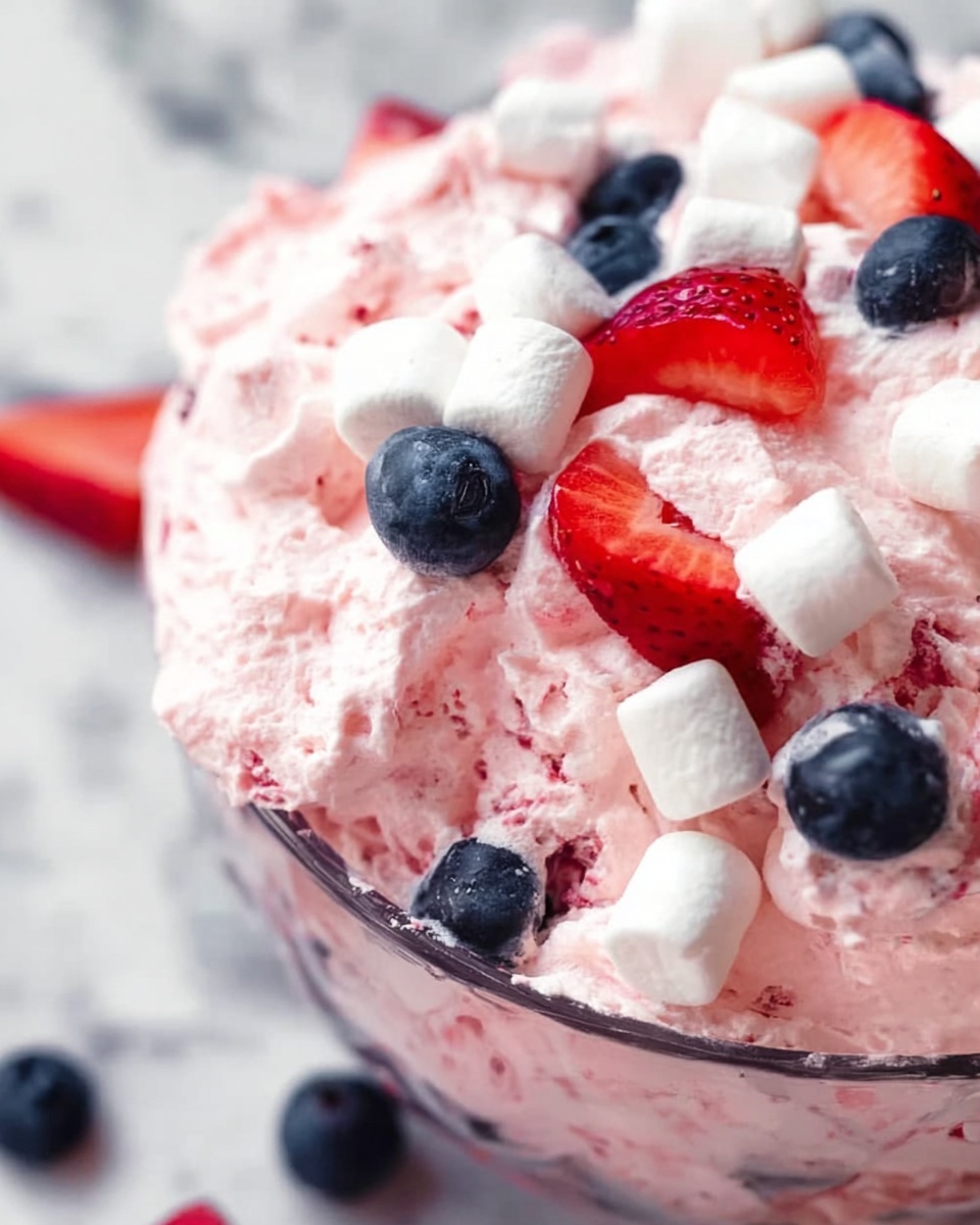 A close-up of a glass bowl filled with fluffy pink whipped dessert, looking creamy and light. The dessert is topped with whole blueberries and fresh strawberry slices, all scattered on top, along with several small white marshmallows mixed in. The bowl sits on a white marbled surface. The colors are soft pinks, deep blues, bright reds, and white. A woman's hand is visible holding the bowl. photo taken with an iphone --ar 4:5 --v 7