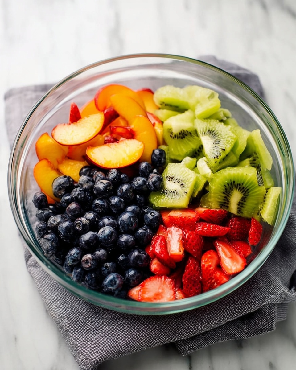 A clear glass bowl sits on a gray cloth on a white marbled surface, filled with four types of fresh fruit arranged in separate sections. One section holds dark blue blueberries with a smooth texture, next to brightly colored orange peach slices with a soft, slightly fuzzy look. Another section contains light green kiwi pieces with visible black seeds and a juicy texture, while the last section features red strawberry slices showing their juicy insides. The fruits create a colorful and fresh mix inside the bowl. photo taken with an iphone --ar 4:5 --v 7