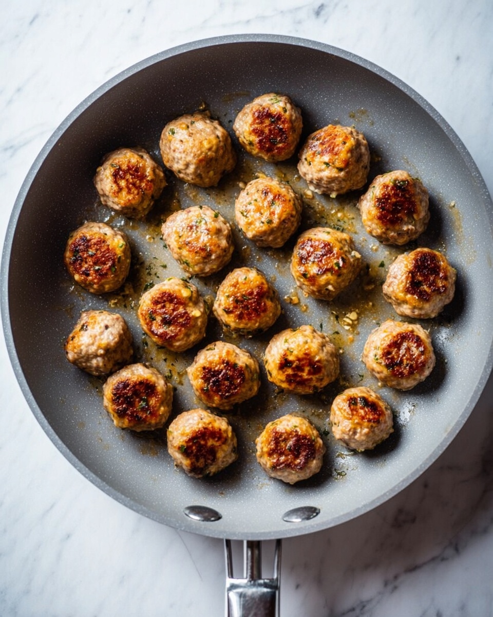A gray frying pan filled with about 18 small, round meatballs, each with a golden brown crust on top. The meatballs are spread evenly across the pan, showing a mix of light and darker brown colors with some specks of herbs visible. The pan is sitting on a white marbled surface. Photo taken with an iphone --ar 4:5 --v 7