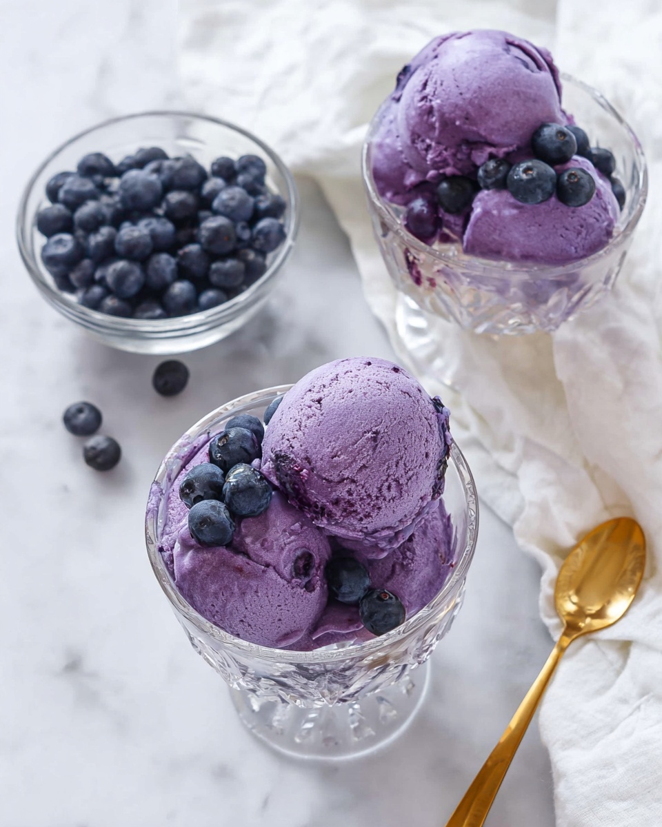 The image shows two clear glass dessert bowls filled with three purple scoops of blueberry ice cream each. The ice cream has a smooth and creamy texture with deeper purple areas. Each bowl is decorated with several fresh, dark blue blueberries placed on top and around the ice cream scoops. There is a smaller clear glass bowl to the side filled with more fresh blueberries. The bowls are placed on a white marbled surface with a white cloth near one bowl and a shiny gold spoon beside it. photo taken with an iphone --ar 4:5 --v 7