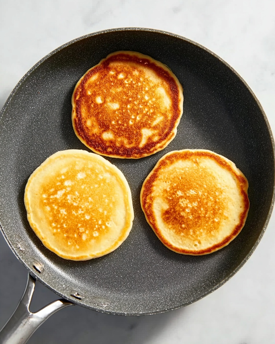 A close-up image shows three small, round pancakes cooking in a dark gray non-stick pan. The pancakes have a golden-brown top layer with slightly uneven edges and small bubbles on their light yellow surface. One pancake at the top has a slightly darker, more toasted upper side, while the other two pancakes below are lighter but still golden. The pan is set on a white marbled surface. The handle of the pan is metallic and partly visible. Photo taken with an iphone --ar 4:5 --v 7