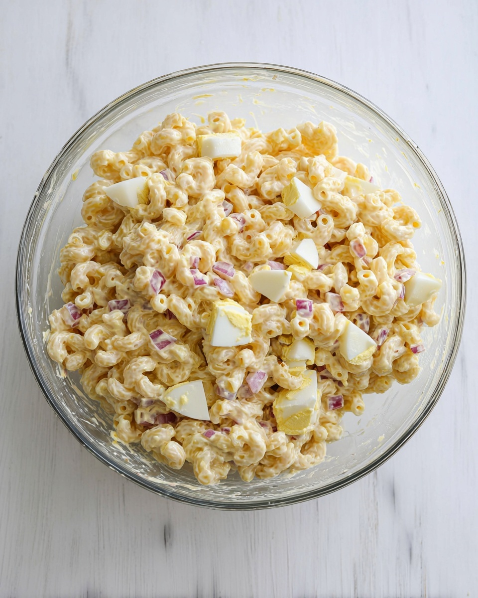 A clear glass bowl filled with creamy macaroni salad sits on a white marbled surface. The salad has three main layers: pale yellow macaroni pasta with a smooth texture, diced white hard-boiled eggs, and small pieces of red onion scattered evenly throughout. The creamy dressing coats all ingredients, giving a light shine and holding the salad together in a thick mixture. The bowl is about half full, showing smears of the dressing on its clear sides. Photo taken with an iphone --ar 4:5 --v 7