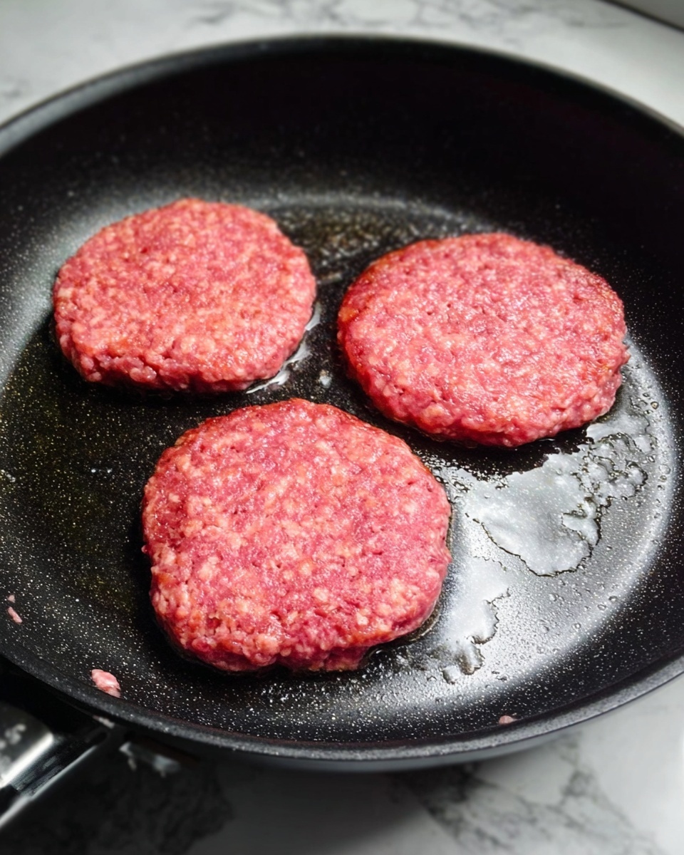 Croissant Smash Burger Recipe 4 The image shows three raw ground meat patties cooking in a black frying pan. The patties are round and have a pinkish-red color with a rough, uneven texture. The pan has some shiny oil spots around the patties. The background is a white marbled surface. Photo taken with an iphone --ar 4:5 --v 7