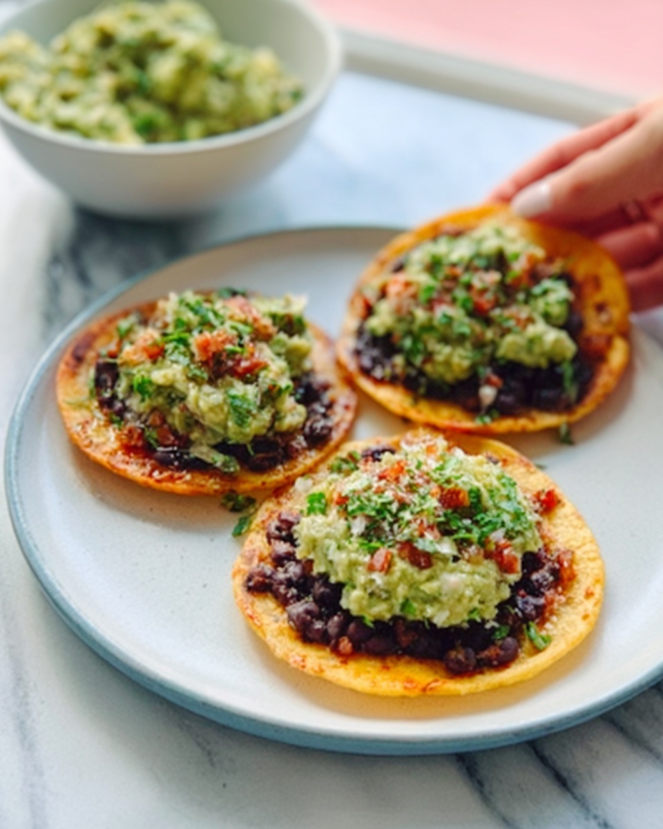 The image shows three small round tostadas on a white plate. Each tostada has a crispy golden base layer. On top of this base, there is a spread of mashed green avocado mixed with small bits of red tomatoes and sprinkled with green herbs. One tostada has some black beans spread under the avocado mix, adding a darker texture. In the background, there is a white bowl filled with more mashed avocado. The scene is on a white marbled surface, with a woman's hand about to pick up one tostada photo taken with an iphone --ar 4:5 --v 7