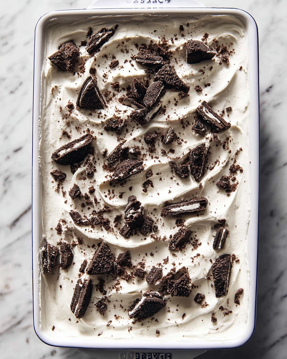 Easy No Bake Oreo Ice Cream Dessert Recipe 5 The image shows a white rectangular dish filled with a thick white creamy layer spread evenly across the surface, with slight wave-like textures. Scattered over the cream are many pieces of broken dark chocolate sandwich cookies with white cream filling, creating a contrast of dark brown and white colors across the top. The dish sits on a white marbled surface, and the photo is taken from above, highlighting the textures and colors clearly. Photo taken with an iphone --ar 4:5 --v 7