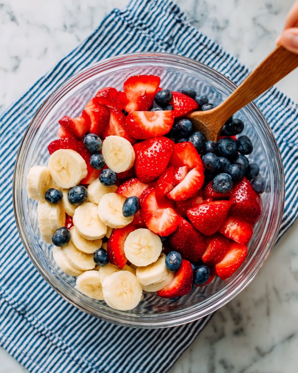 The image shows a clear round bowl filled with fresh fruit salad. The bowl is on a white marbled surface with a blue and white striped cloth underneath. Inside the bowl, the bottom is layered with round pieces of banana that are light yellow and smooth. On top of the banana, there are small, round dark blue blueberries scattered evenly. The top layer has bright red strawberries, some whole and some sliced in halves, with their shiny texture visible. A woman's hand is holding a wooden spoon that is partially in the bowl, positioned on the right side. Photo taken with an iphone --ar 4:5 --v 7