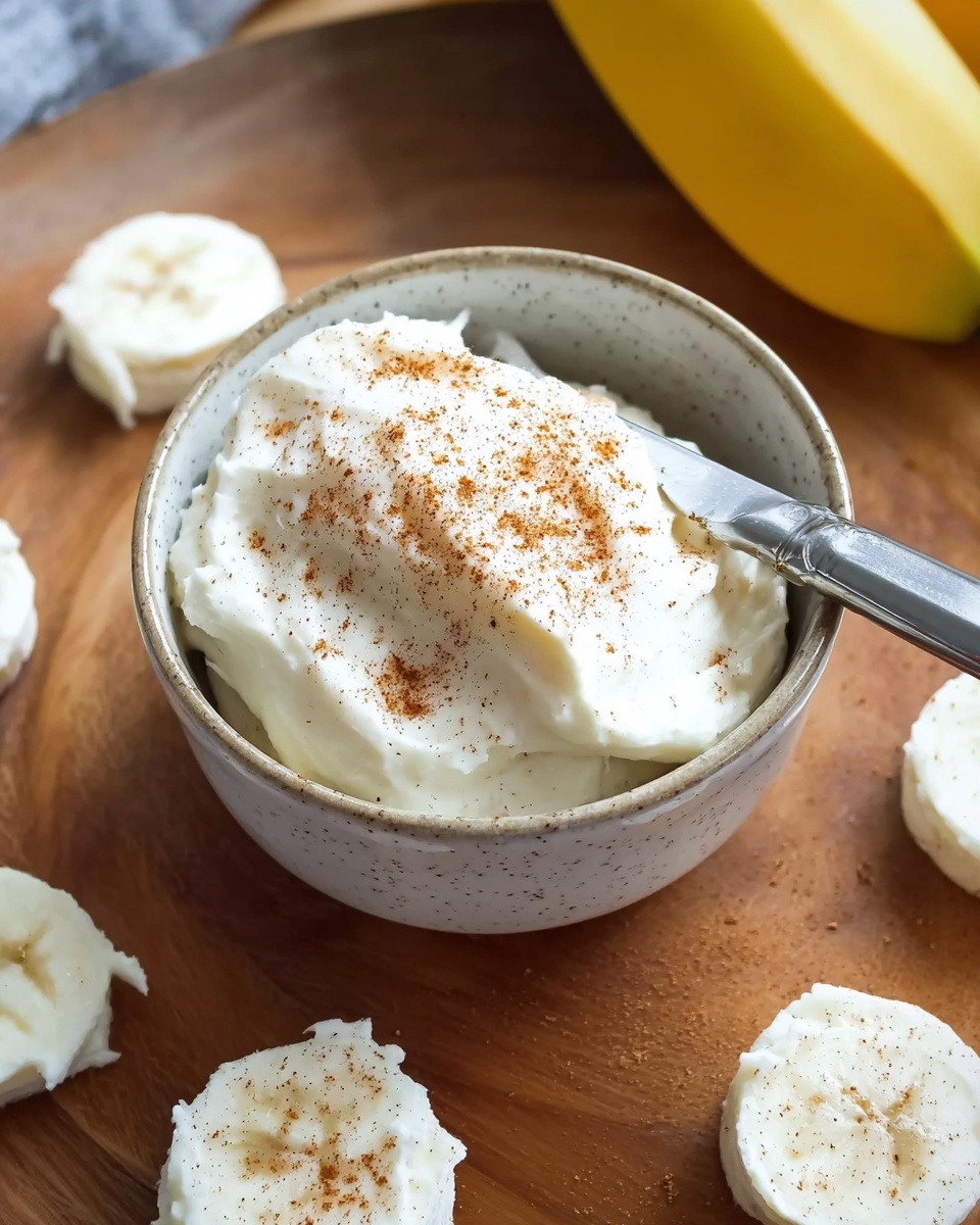A small white speckled bowl holds a thick, creamy white spread lightly dusted with brown cinnamon on top, with a silver knife resting diagonally inside the bowl. Around the bowl on a wooden surface, there are several dollops of the white spread shaped in small circles, and in the background, a yellow banana peels slightly visible. Photo taken with an iphone --ar 4:5 --v 7