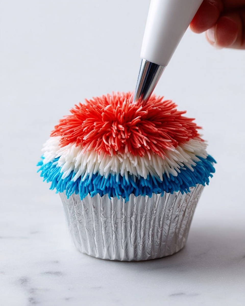 A single cupcake in a silver foil wrapper sits on a white marbled surface. The cupcake is decorated with three layers of thin, long icing strands that look like fur: a red layer on top center, a white layer underneath red covering the middle area, and a blue layer covering the bottom edge all around. A piping bag is adding more red icing on the top center, showing a woman's hand holding the tip. The background is simple and white, focusing fully on the colorful textures of the cupcake. photo taken with an iphone --ar 4:5 --v 7