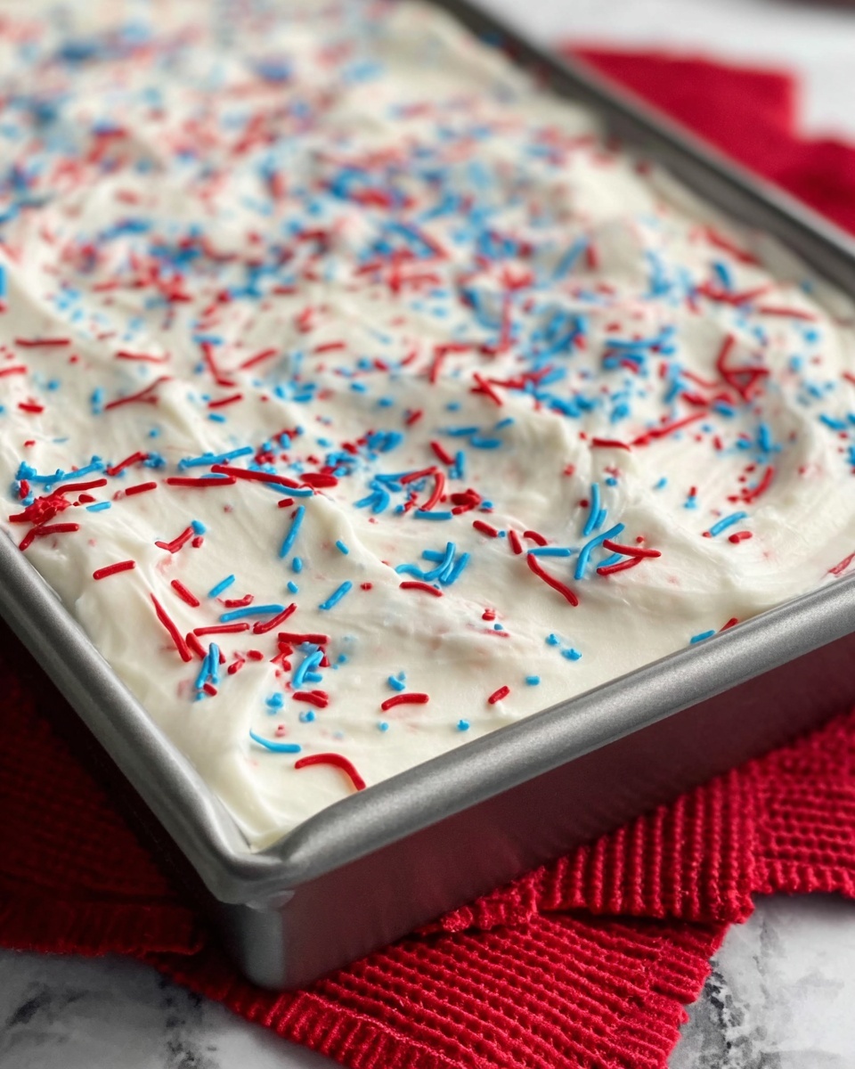 The image shows a close-up of a rectangular metal pan filled with a thick, white creamy layer, spread unevenly across the top. Scattered on the white layer are red and blue sprinkles, along with thin swirls of red and blue, creating a playful, colorful pattern on the smooth surface. The pan is placed on a white marbled surface with a red textured cloth underneath it. The angle of the photo shows a detailed texture of the cream and decorations, emphasizing the contrast of colors and soft, fluffy texture of the topping. Photo taken with an iphone --ar 4:5 --v 7