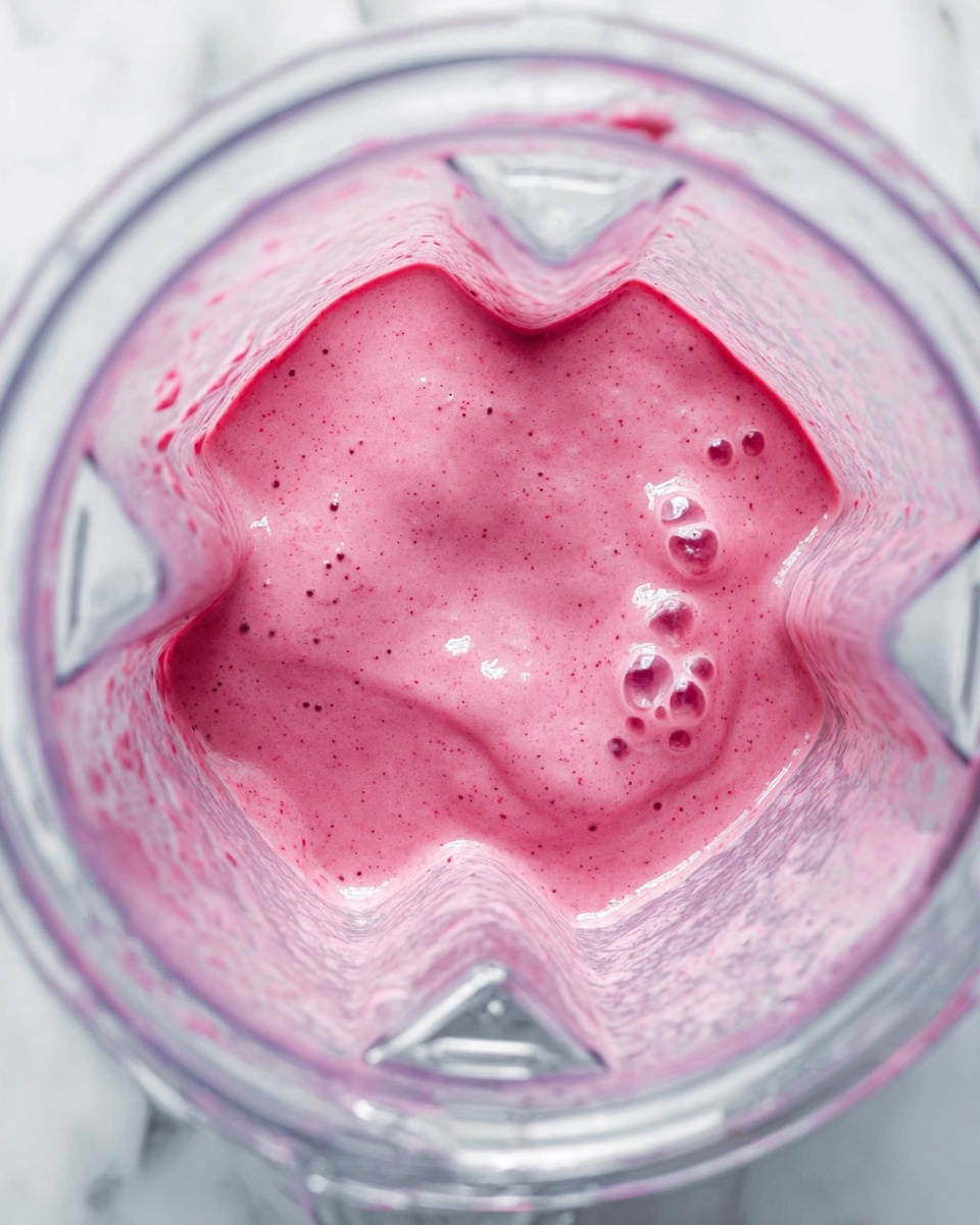 The image shows a close-up top view of a blender container filled with a thick, pink smoothie mixture. The smoothie has a creamy texture with small dark speckles and bubbles scattered on the surface, giving a fresh, blended appearance. The blender container is clear with visible interior ridges, and the background surface is a white marbled texture. Photo taken with an iphone --ar 4:5 --v 7
