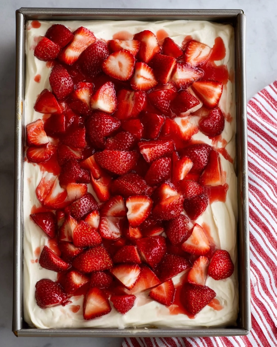 This image shows a rectangular metal pan filled with a dessert made of two main layers. The bottom layer is a thick, creamy white base with a smooth and soft texture, spread evenly to fill the pan. On top of this creamy layer, there is a generous layer of bright red strawberries, some whole, some sliced in half, scattered irregularly across the surface. The strawberries are shiny with juice, adding a fresh and vibrant contrast to the white base. The pan is placed on a white marbled surface, and a striped red-and-white cloth is partially visible on the right side. Photo taken with an iphone --ar 4:5 --v 7