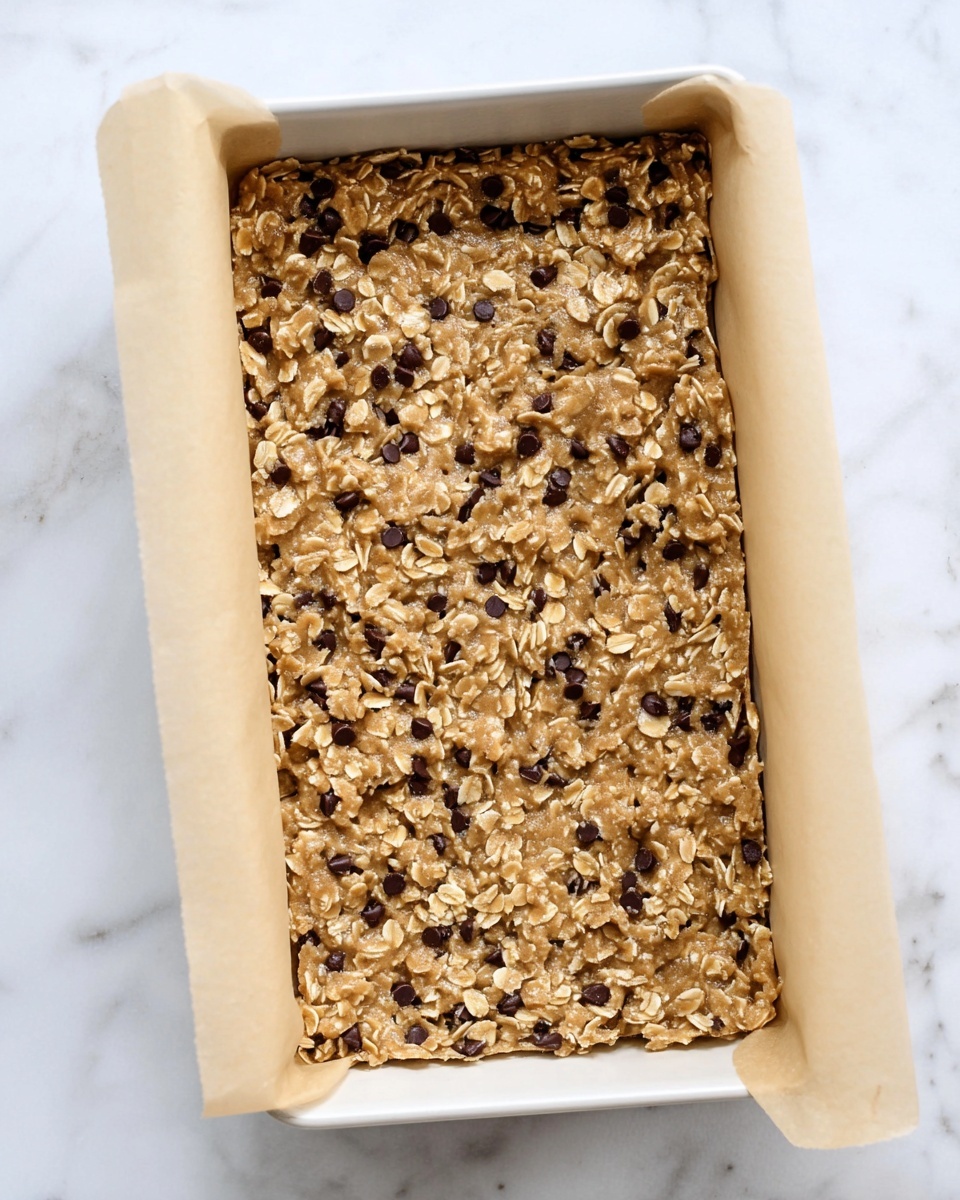 The image shows a thick layer of raw oat cookie dough mixed with small dark chocolate chips spread evenly in a rectangular white tray lined with beige parchment paper. The dough has a rough texture with visible oat flakes throughout. The tray is placed on a smooth white marbled surface. photo taken with an iphone --ar 4:5 --v 7