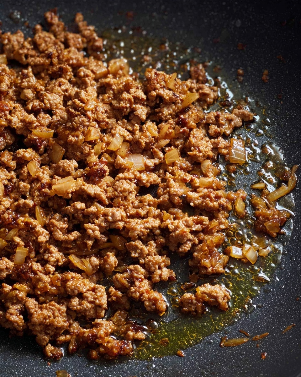 The image shows cooked ground meat with small pieces of onions in a black pan. The meat is brown with some golden edges, and there is shiny oil around the meat, creating a glistening effect. The texture of the meat is crumbly and uneven, with some parts clumped together. The pan surface is dark and smooth, contrasting with the rich brown color of the meat. The photo taken with an iphone --ar 4:5 --v 7