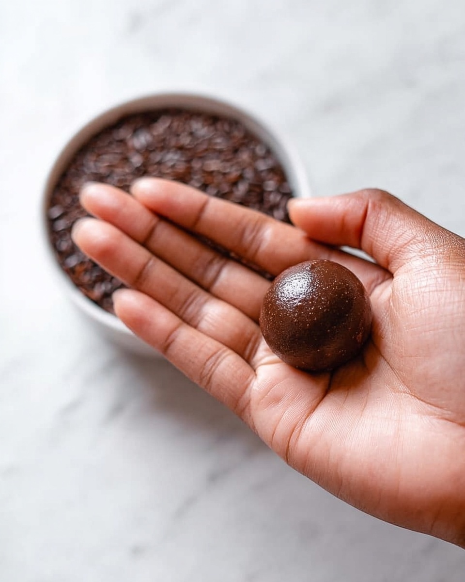 Brigadeiro Recipe 4 A woman's hand is held open with the palm facing up, holding a small round ball of smooth, dark brown chocolate dough. The texture of the ball looks soft and shiny. Behind the hand, there is a white bowl filled with dark brown grains or seeds, placed on a white marbled surface. The focus is mainly on the hand and the chocolate ball. photo taken with an iphone --ar 4:5 --v 7