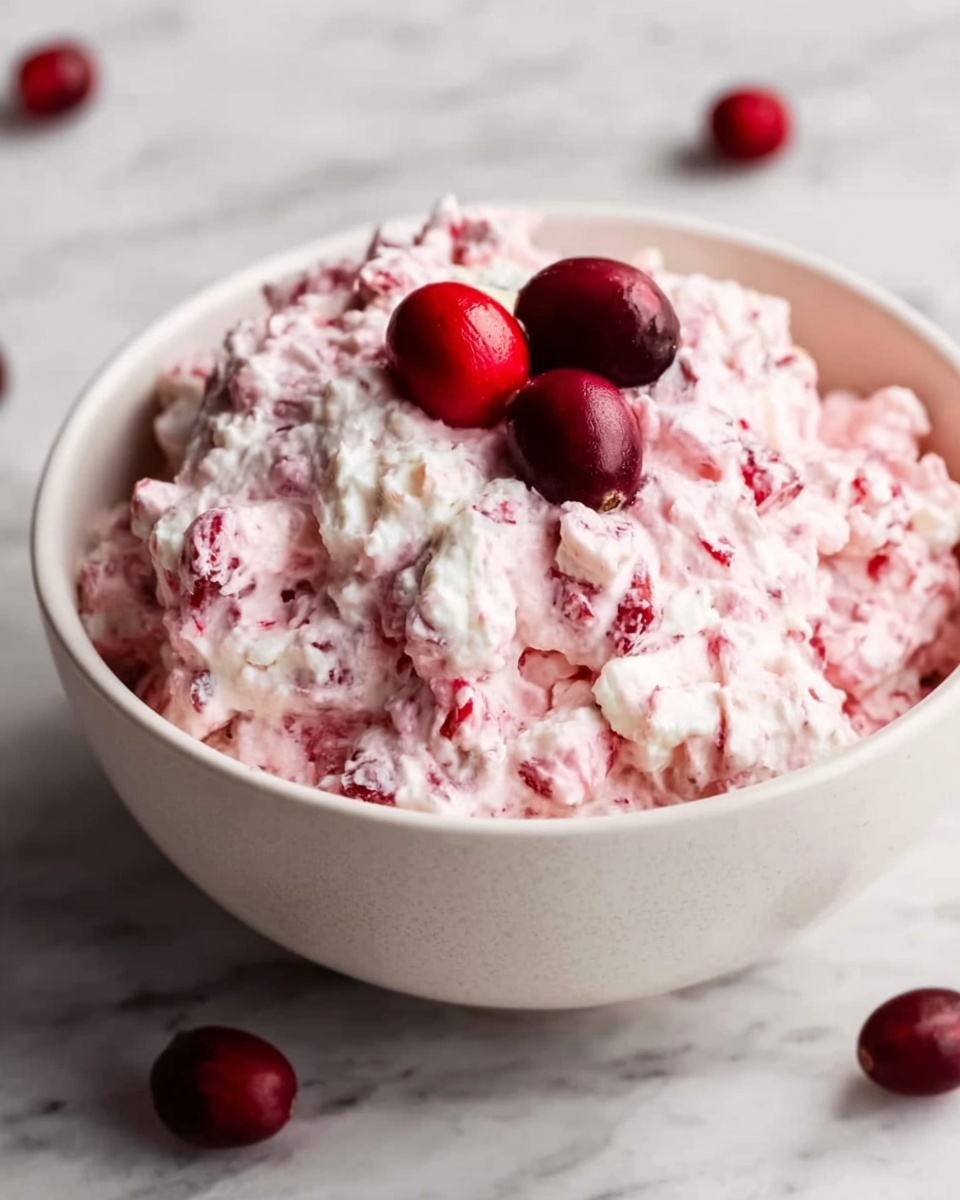 A white bowl filled with a creamy, thick pink and white mixture that looks soft and fluffy, with visible small chunks inside. On top of the mixture, there are three whole shiny deep red cranberries placed together. The bowl sits on a white marbled surface with a few red cranberries scattered around. The texture of the mixture is uneven and slightly lumpy, showing bits of red mixed well into the cream. photo taken with an iphone --ar 4:5 --v 7