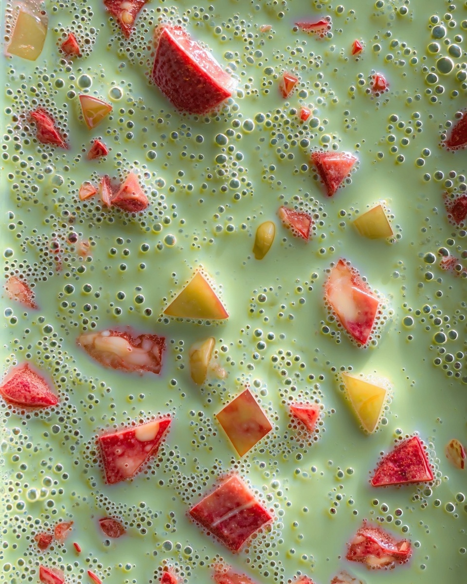 The image shows a close-up of a creamy green liquid base with many small bubbles on the surface. Mixed within the green base are small colorful pieces, including red and pink chunks, some light orange bits, and a few translucent pale blue shards, scattered evenly throughout. The texture looks smooth and slightly thick, with the bubbles giving it a fresh and lively look. The background is a white marbled texture. Photo taken with an iphone --ar 4:5 --v 7