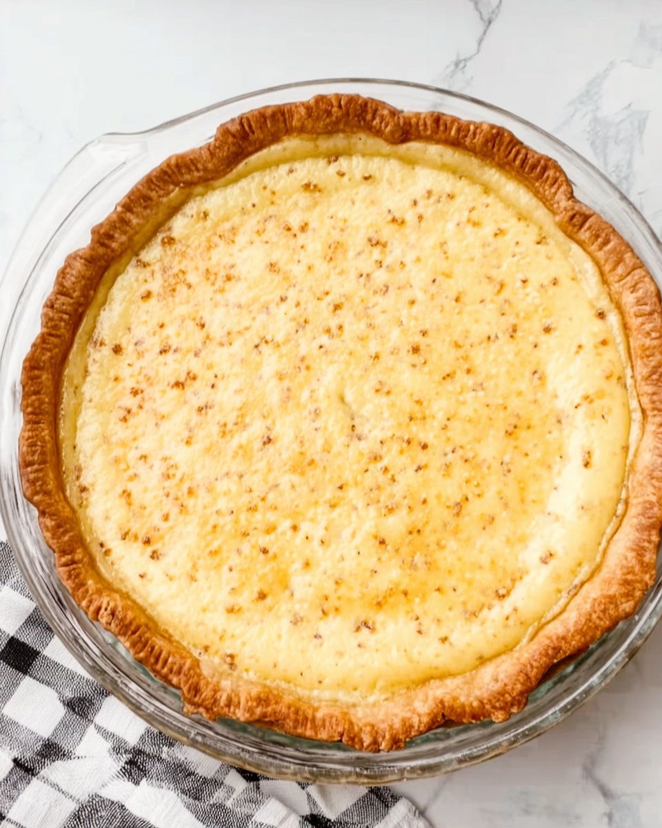 Easy Custard Pie Recipe 4 The image shows a whole pie in a clear glass pie dish with a golden brown crust that is slightly crimped around the edges. The filling is a smooth, creamy light yellow color with some small darker spots, indicating a baked surface. The pie is set on a white marbled surface with a black and white checkered cloth partially visible nearby. The overall look is warm and inviting, with a clean and simple presentation. Photo taken with an iphone --ar 4:5 --v 7