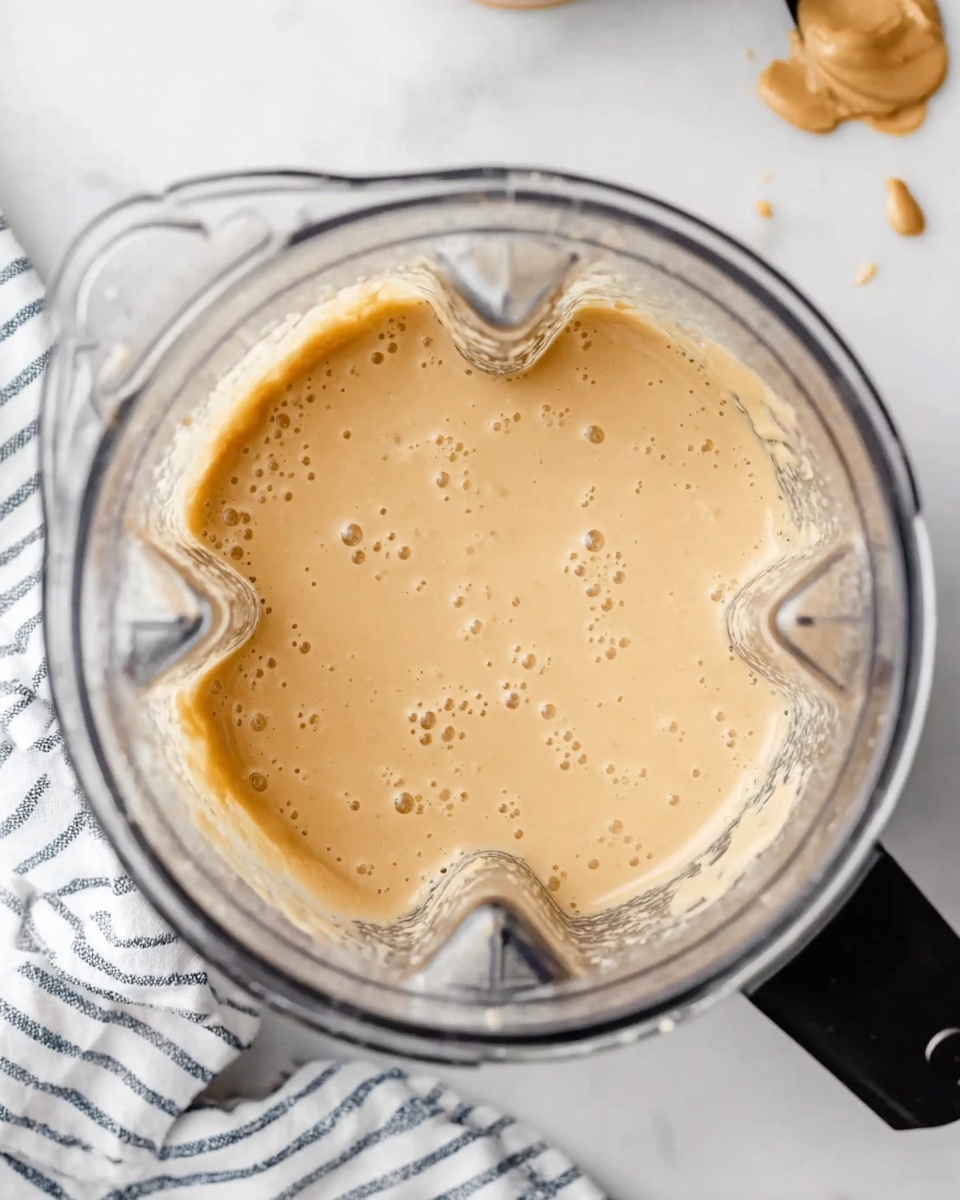 The image shows a clear blender container filled with a creamy, light beige mixture with a smooth but slightly lumpy texture. Tiny bubbles and small lumps can be seen throughout the creamy layer inside the blender. The blender is placed on a white marbled surface, with a black spatula smeared with peanut butter lying nearby, and a towel with white stripes partially visible under the blender. A woman's hand is not visible in the image. photo taken with an iphone --ar 4:5 --v 7