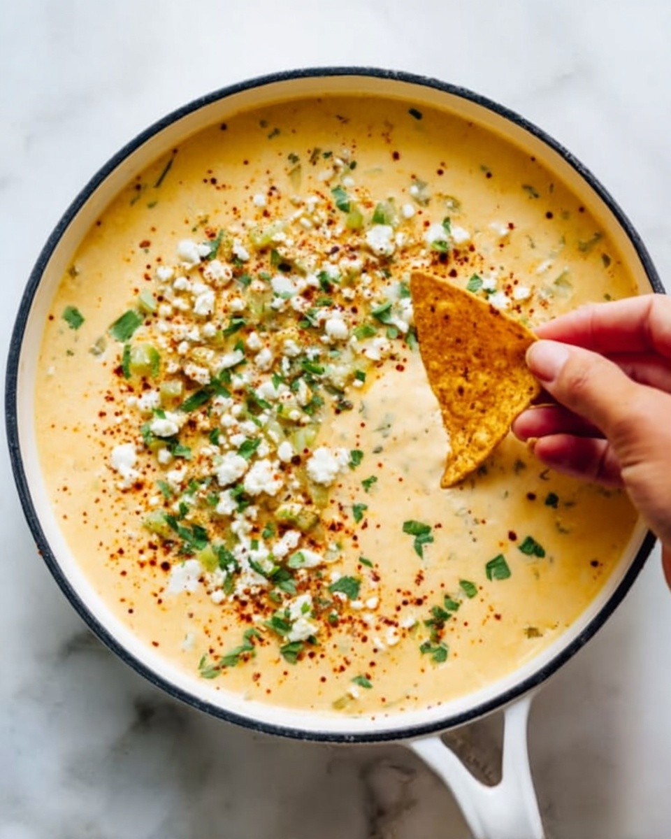 A woman's hand is dipping a triangular light yellow tortilla chip into a creamy, smooth, light beige cheese sauce. Around the sauce, there is a topping layer with small golden-brown grilled corn kernels, white cheese crumbles, and scattered green herb leaves, all on a white marbled surface. The image is close-up with soft lighting highlighting the textures. photo taken with an iphone --ar 4:5 --v 7