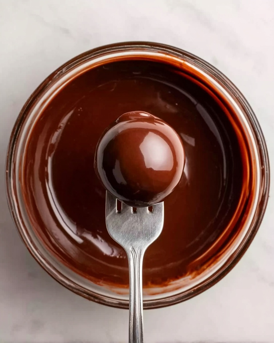 A clear glass bowl filled with smooth, shiny dark chocolate. In the middle of the bowl, a fork holds a round chocolate-covered ball with a glossy finish. The chocolate coating looks thick and even, reflecting light gently. The bowl sits on a white marbled surface. photo taken with an iphone --ar 4:5 --v 7