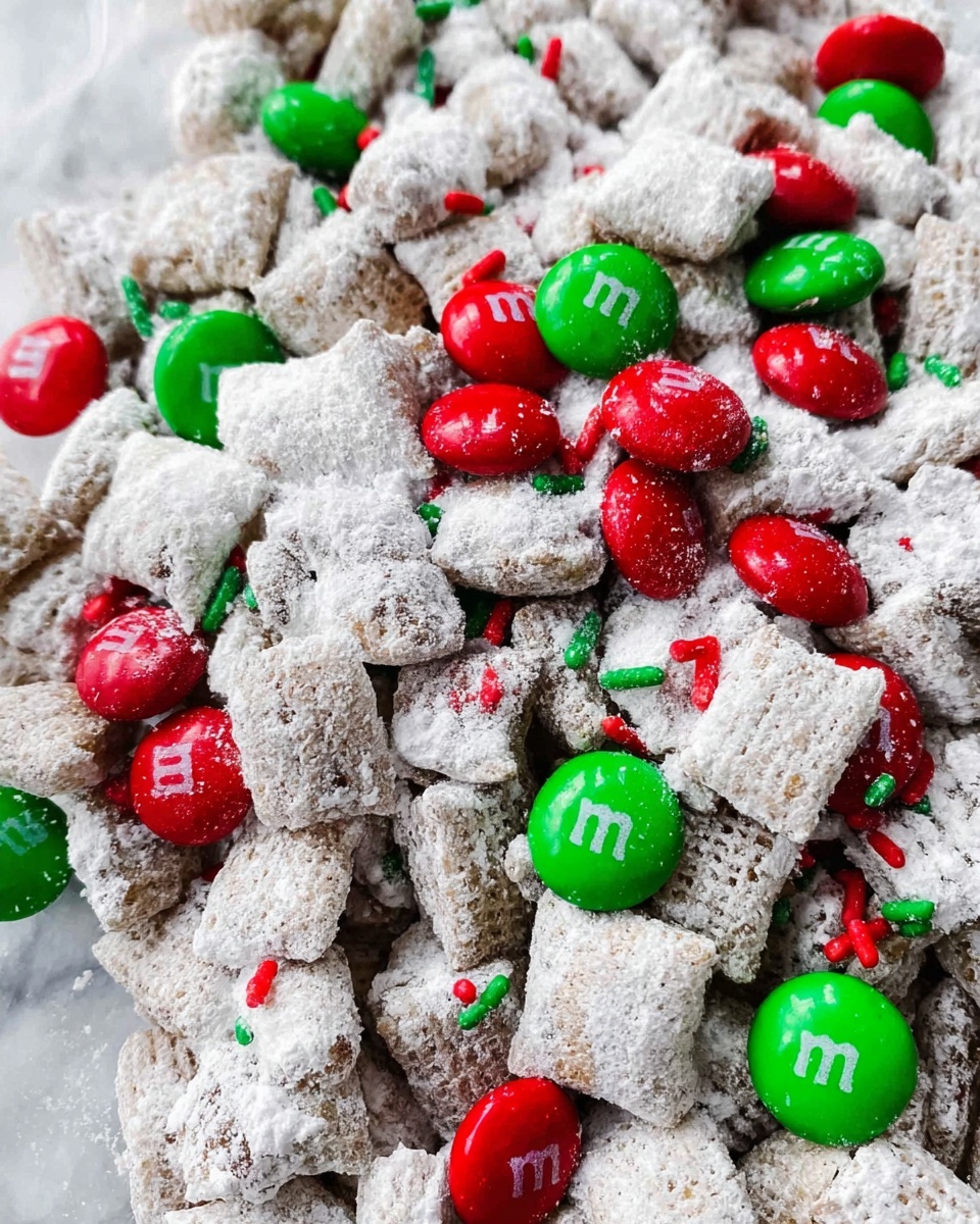 A white bowl filled with a snack mix that has multiple layers: the bottom layer is covered with clusters of light brown pieces coated thickly in white powdered sugar, giving them a rough texture; mixed throughout are shiny red and green candy-coated chocolates that are smooth and round, some with a small white letter