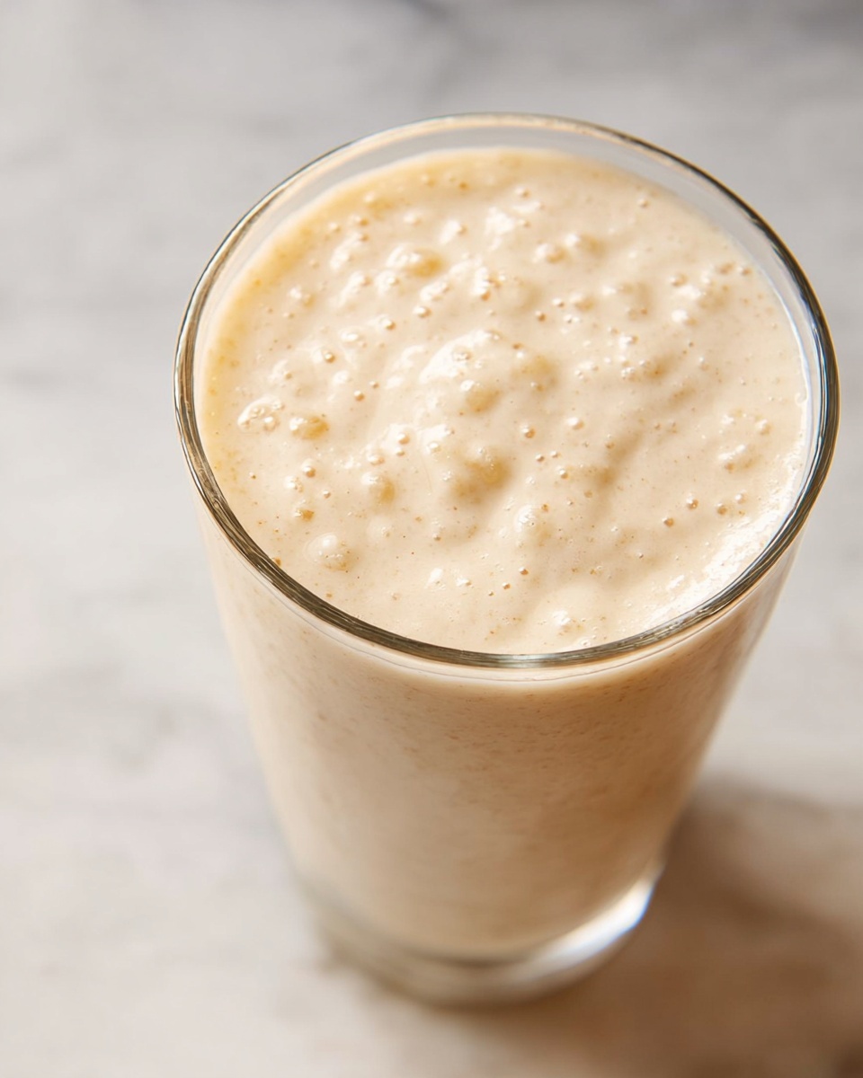 A close-up view of a tall clear glass filled with a thick, creamy beige smoothie. The smoothie has a frothy texture on top with small bubbles and soft peaks, showing a smooth and slightly airy consistency. The glass sits on a white marbled surface, and the background is softly blurred, keeping the focus on the drink's surface details. photo taken with an iphone --ar 4:5 --v 7