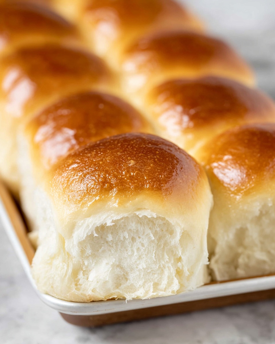 A close-up view of a white tray filled with nine soft bread rolls in three rows. One roll at the front left has been pulled up, showing its fluffy white inside and a golden brown top. The rolls have smooth, shiny textures on top with warm, light brown colors fading softly into the white fluffy sides and inner crumb. The tray sits on a white marbled surface in a bright light setting. photo taken with an iphone --ar 4:5 --v 7