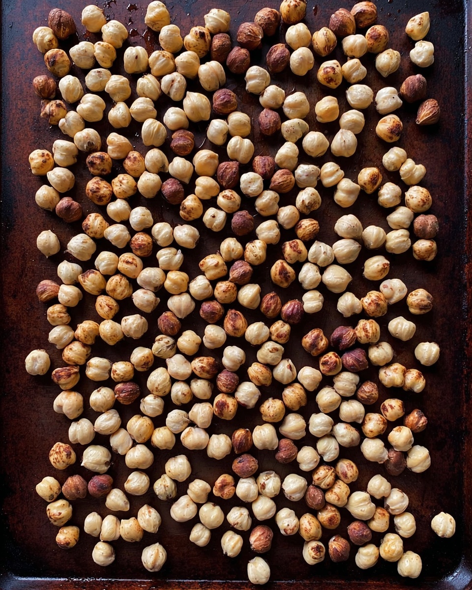 A dark metal baking sheet with a flat surface is spread evenly with a single layer of small, round hazelnuts. The hazelnuts vary in colors from pale beige to light brown, with some showing darker brown spots from roasting. The nuts are scattered in a way that covers nearly the whole baking sheet with little empty space, and the background is not visible beneath them. The image is sharp and focused, showing the texture and slight shine on the surface of each hazelnut. photo taken with an iphone --ar 4:5 --v 7
