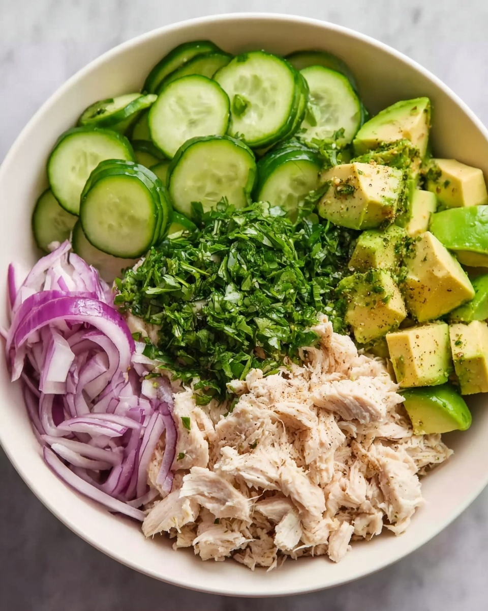A white bowl on a white marbled surface holds a fresh salad arranged in distinct sections. On one side are thick, green cucumber slices with some seasoning on top. Next to them is a pile of finely chopped green herbs, bright and fresh, placed in the center of the bowl. To the right of the herbs, there is light-colored shredded chicken seasoned with black pepper. Toward the bottom left, thin rings of purple onion add color contrast. Lastly, there are small cubes of green avocado, also sprinkled with seasoning. The whole dish looks healthy and colorful. Photo taken with an iphone --ar 4:5 --v 7