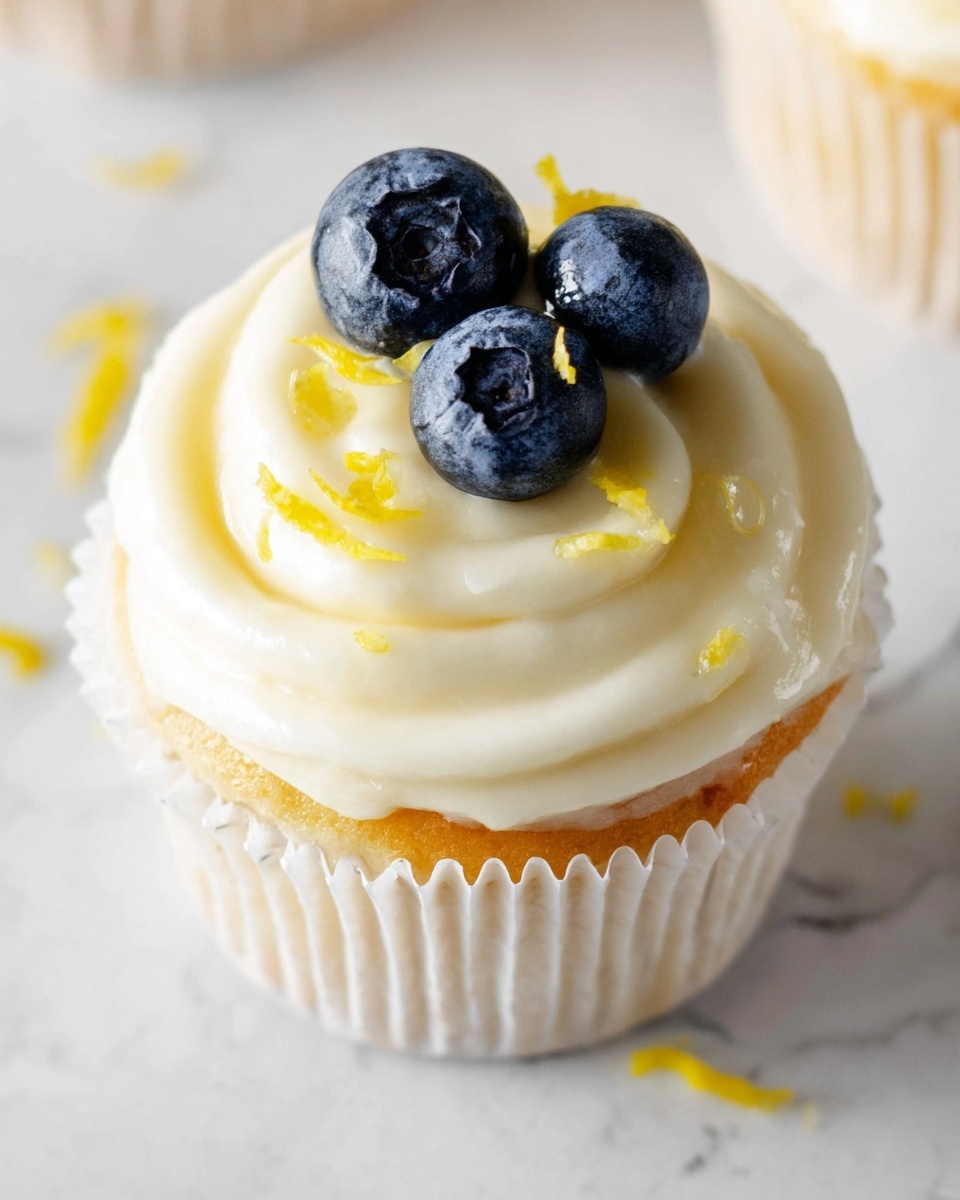 The image shows several cupcakes arranged on a metal cooling rack placed on a white marbled surface. Each cupcake has a golden brown base with a smooth white paper liner. On top, there is a thick, swirled layer of creamy white frosting, which is decorated with three fresh blueberries in the center and small pieces of yellow lemon zest sprinkled around. The texture of the frosting looks soft and smooth, with a gentle swirl pattern standing out clearly. photo taken with an iphone --ar 4:5 --v 7