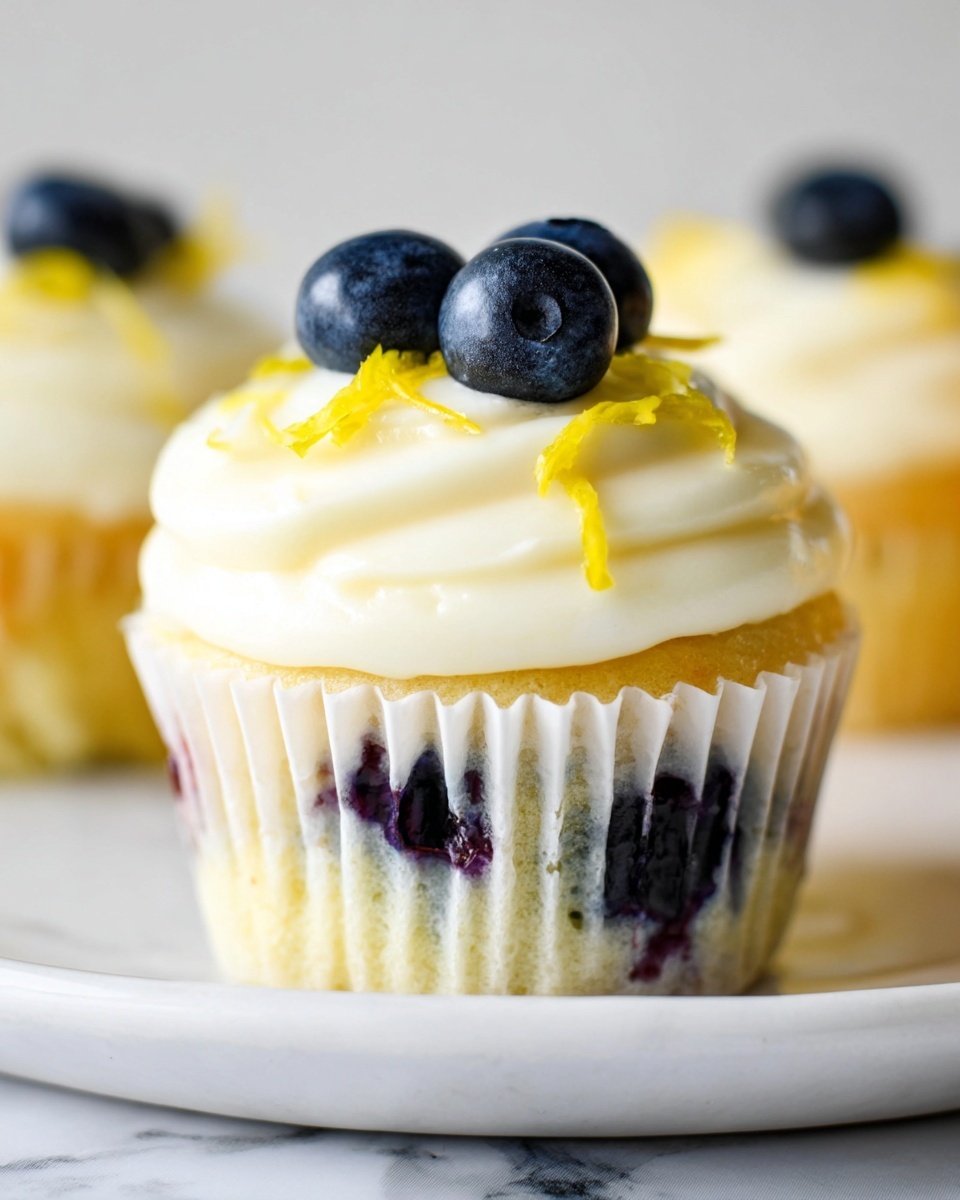 A single cupcake with a light golden base visible through white paper liner, topped with a thick swirl of smooth creamy white frosting, decorated with three plump dark blue blueberries placed at the center and thin bright yellow lemon zest strips scattered gently around. The cupcake sits on a white marbled surface with a soft natural light highlighting the fresh texture and colors of the frosting and fruit, photo taken with an iphone --ar 4:5 --v 7