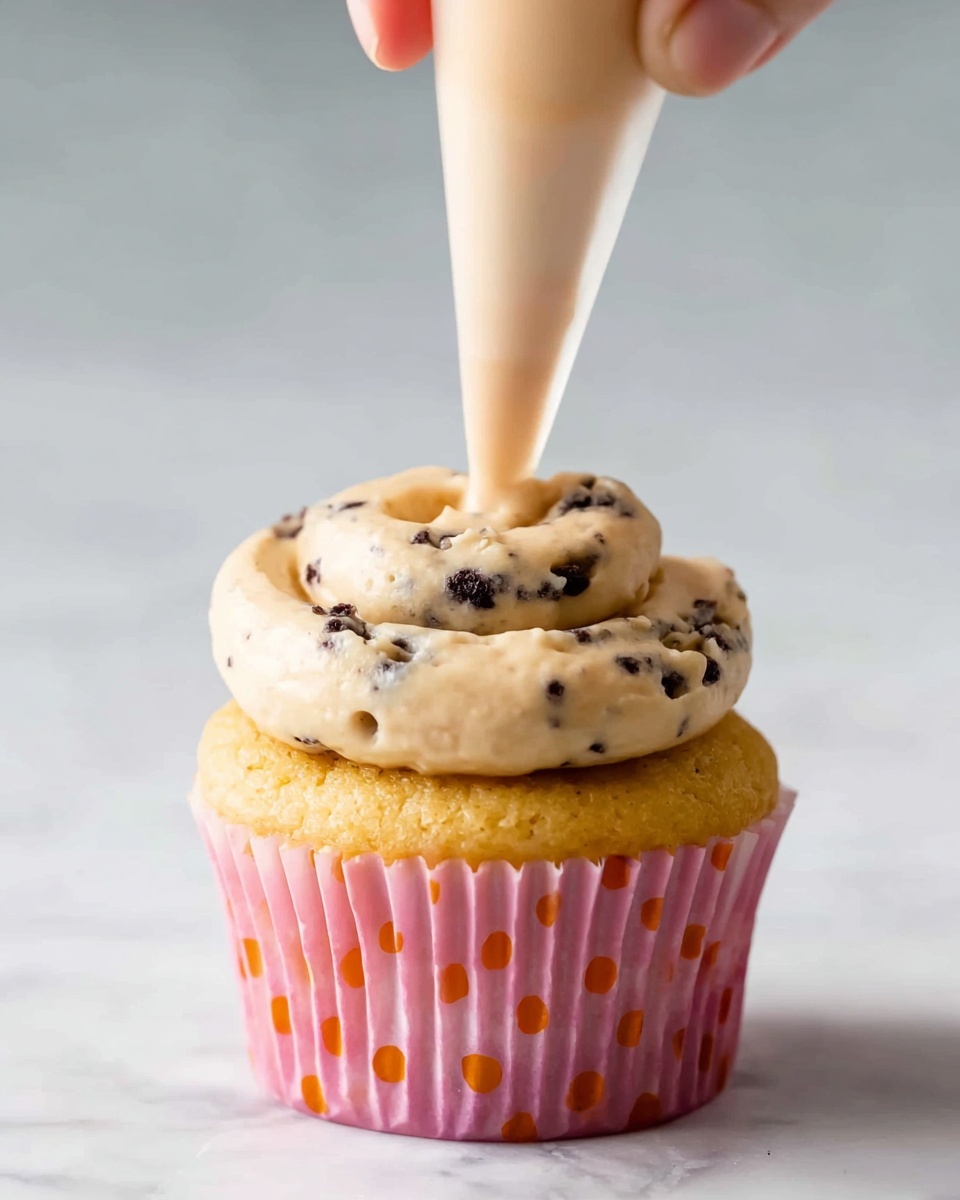 A close-up image shows a single cupcake in a pink cupcake liner with small orange polka dots. The cupcake is light golden yellow and sits on a white marbled surface. On top of the cupcake, a swirl of light beige frosting with tiny dark chocolate bits is being piped from a clear plastic piping bag held by a woman's hand. The frosting is thick with a smooth but textured surface and is forming a single round layer on the cupcake top. photo taken with an iphone --ar 4:5 --v 7