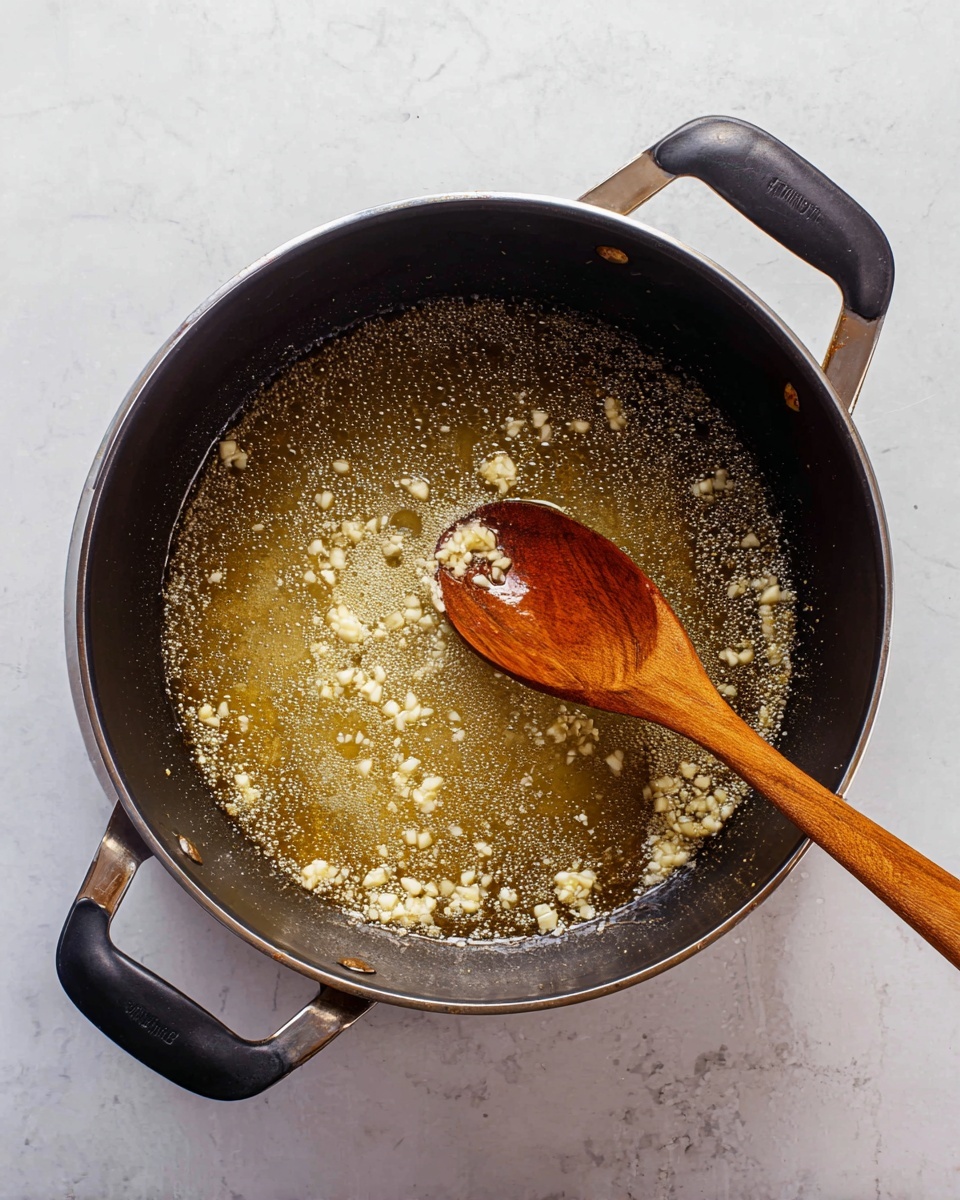 The image shows a deep pan with two black handles on each side, sitting on a white marbled surface. Inside the pan, there is a layer of bubbling golden oil with small pieces of light beige chopped garlic spread evenly throughout. A wooden spoon with a rich brown, smooth texture rests diagonally inside the pan, partly submerged in the oil. The scene is simple, focused on the garlic cooking in the oil under warm light. photo taken with an iphone --ar 4:5 --v 7