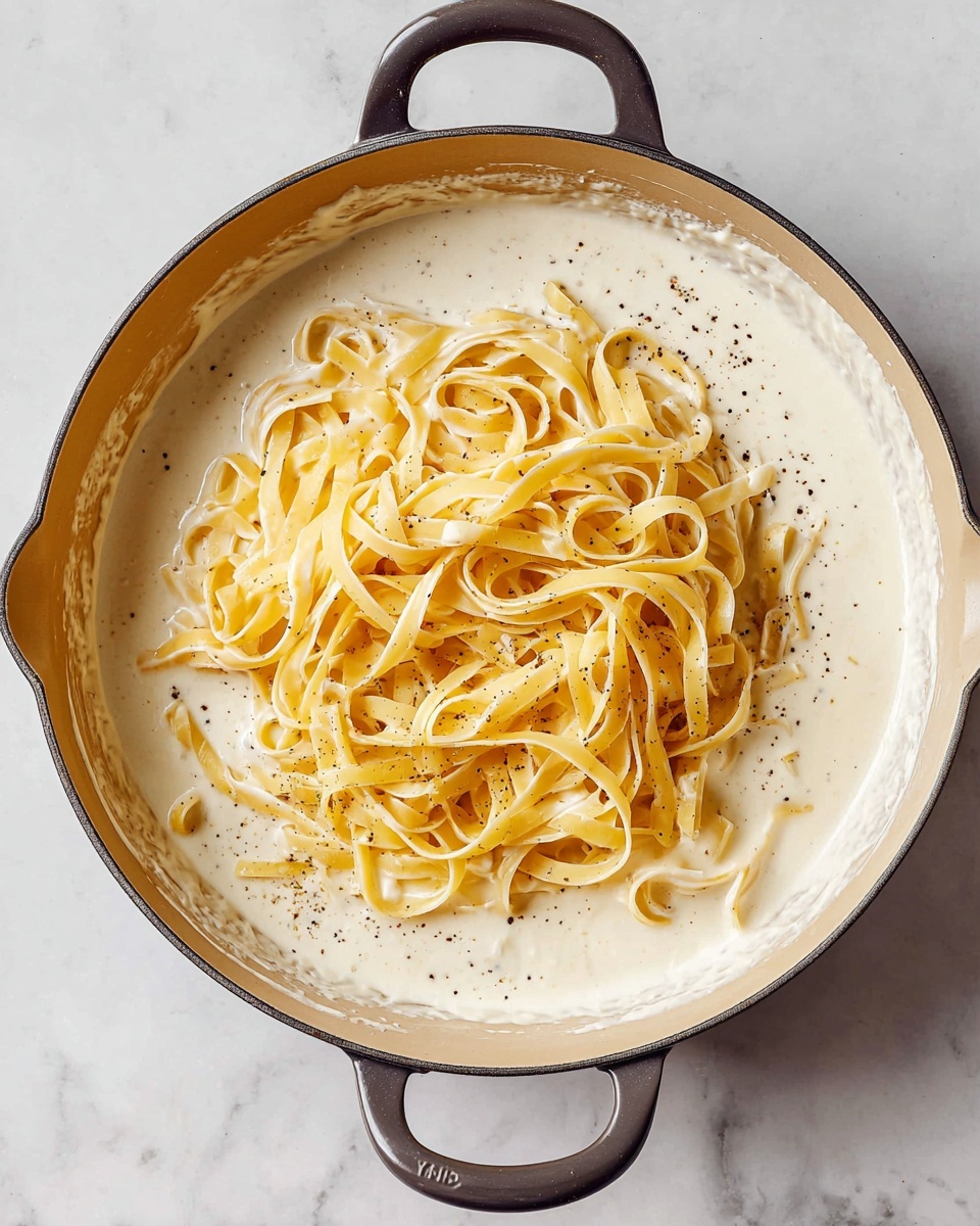 Inside a beige pan with two dark handles, there is a pile of cooked pale yellow fettuccine pasta resting on a creamy white sauce that fills the rest of the pan. The sauce has a smooth and thick texture with small specks of black pepper, and it covers the entire base of the pan except where the pasta sits, creating a contrast between the creamy white and soft yellow colors. The pan is placed on a white marbled surface. photo taken with an iphone --ar 4:5 --v 7