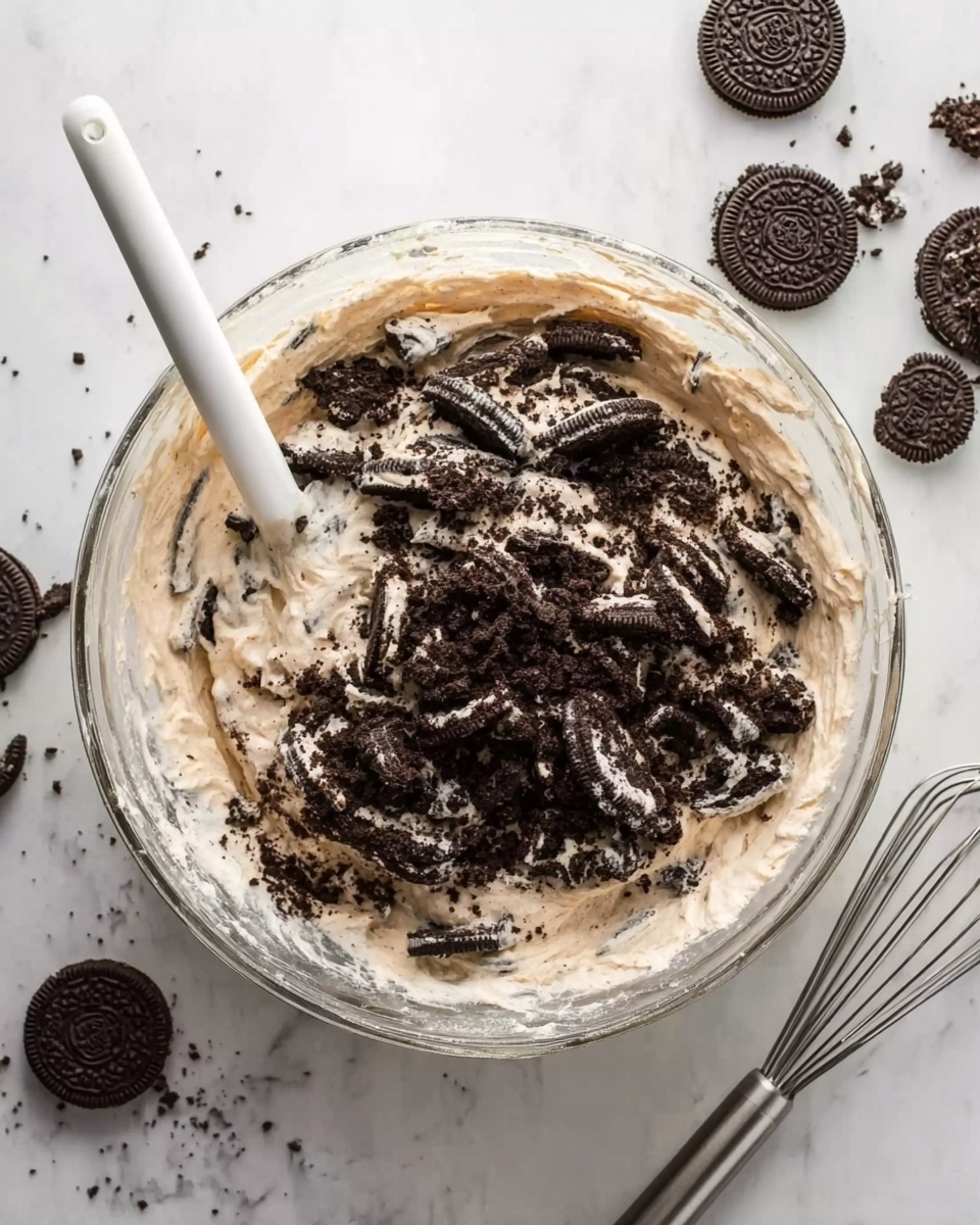 A clear glass bowl sits on a white marbled surface, filled with a creamy, light beige mixture. On top of the mixture, there is a generous layer of crushed dark chocolate sandwich cookies scattered unevenly. A white spatula is partially dipped into the bowl from the left side, spreading the cookie pieces into the creamy base. Around the bowl on the white marbled surface, whole and broken dark chocolate sandwich cookies are scattered, adding texture to the scene. Nearby is a metal whisk resting on the surface to the right. The photo taken with an iphone --ar 4:5 --v 7