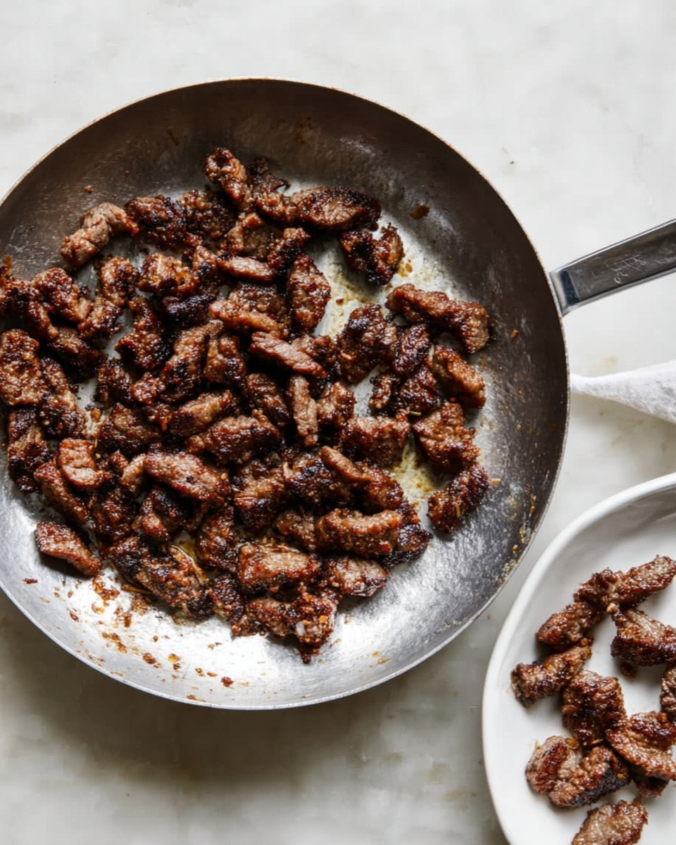 The image shows a silver pan filled with many small, browned, cooked meat pieces that have a slightly crispy texture. The cooked meat pieces are dark brown with some lighter spots and are spread unevenly inside the pan, covering the bottom. Next to the pan, on the right side of the image, there is a white plate with a few pieces of the same cooked meat. The whole scene is set on a white marbled surface. Photo taken with an iphone --ar 4:5 --v 7