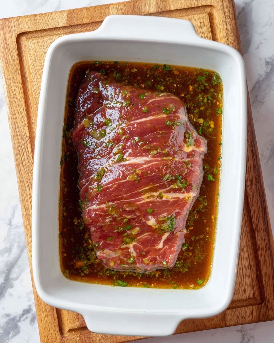 The image shows one large flat piece of raw meat with visible white fat lines, lying flat in the middle of a white rectangular dish. The meat is covered in a shiny brown marinade that fills the dish, with small green herb pieces sprinkled on top and floating in the marinade. The dish is placed on a light wooden board, all set against a white marbled surface. photo taken with an iphone --ar 4:5 --v 7