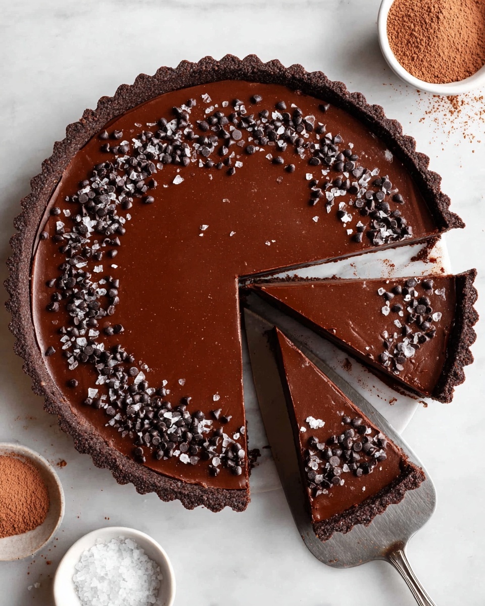 A round chocolate tart with a thick, dark brown crust forms the base layer, holding a smooth, glossy, rich chocolate filling that is evenly spread inside. The top layer is decorated with a ring of small dark chocolate chips and coarse sea salt crystals, adding texture and contrast. Three pie slices are cut and slightly separated from the main tart, with one slice being lifted on a silver spatula showing the clean edges of the filling and crust. The tart is placed on a white marbled surface, and nearby are two small white bowls, one with coarse salt and the other with cocoa powder, adding context to the scene. Photo taken with an iphone --ar 4:5 --v 7