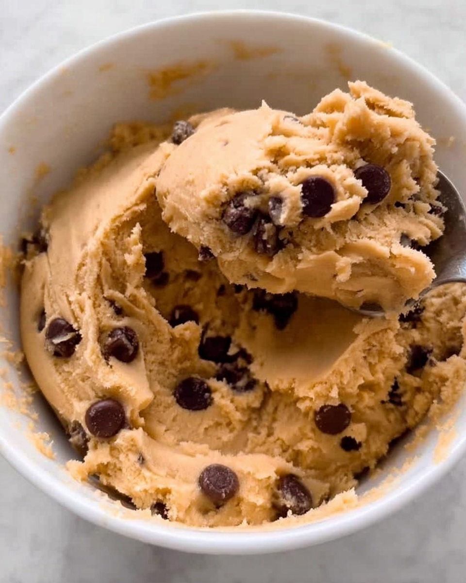 This image shows a white bowl filled with thick, tan cookie dough that has small, dark brown chocolate chips mixed all through it. The dough looks soft and slightly clumpy, with some smooth areas and some rougher, textured spots where the chocolate chips stick out. A spoon has scooped a large amount of dough from the bowl, showing the creamy texture inside. The white bowl contrasts with the cookie dough colors, and the round chocolate chips add dark dots across the tan dough. The background is a white marbled texture. photo taken with an iphone --ar 4:5 --v 7