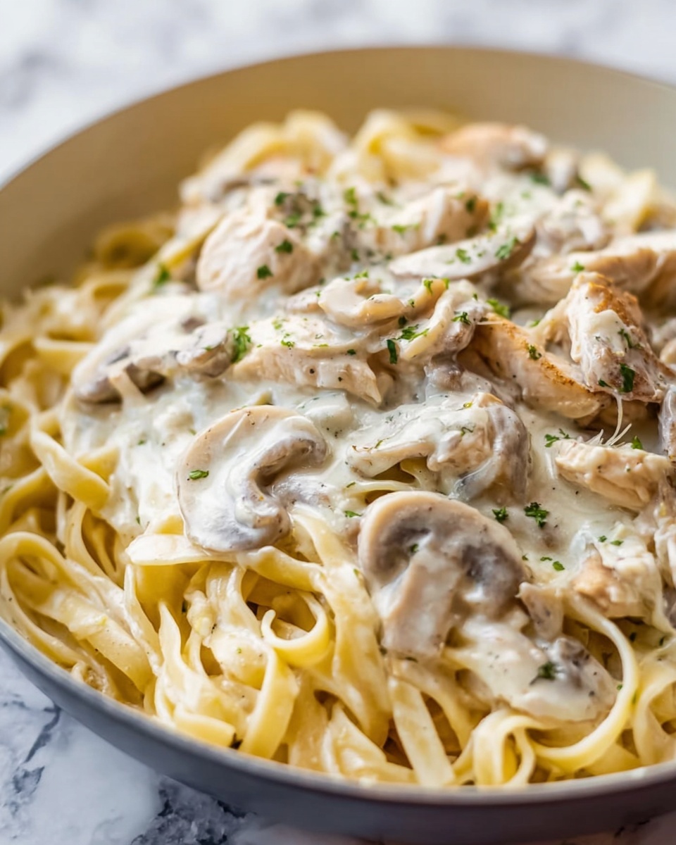 The image shows a close-up of a creamy pasta dish served in a white bowl placed on a white marbled surface. The dish has three main layers visible: the bottom layer consists of long, flat, light beige fettuccine noodles. On top of the noodles is a creamy, white sauce with a smooth texture that coats the pasta evenly. The third layer includes slices of cooked chicken and mushrooms mixed throughout the pasta and sauce, with the chicken pieces being light cream colored and the mushrooms light brown with a soft texture. The dish is sprinkled with small green herb bits, likely parsley, adding a touch of color contrast. Photo taken with an iphone --ar 4:5 --v 7