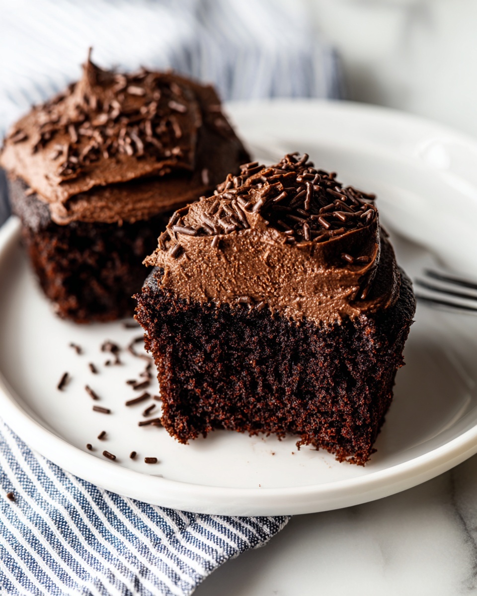 Two pieces of dark brown chocolate cupcake sit on a white plate, with a visible soft and moist texture inside. Each cupcake has one thick layer of smooth, glossy chocolate frosting on top that is slightly textured with small peaks. Dark brown chocolate sprinkles are scattered on the frosting and around the plate. The plate rests on a white marbled surface, with a striped blue and white cloth to the side. Photo taken with an iphone --ar 4:5 --v 7