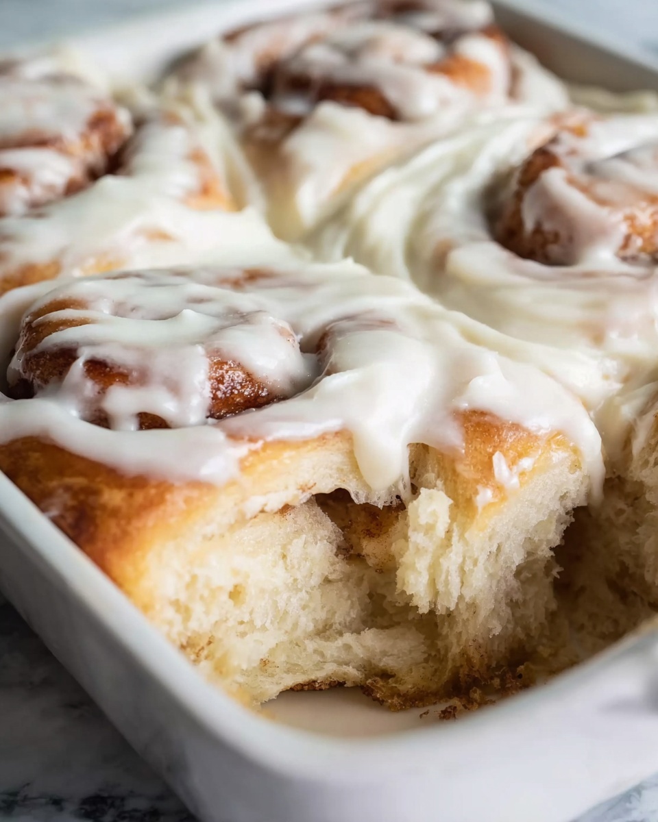 The image shows soft cinnamon rolls in a white dish, each roll covered with a thick layer of white cream cheese icing. The rolls are light brown with a slightly golden crust, and the icing is smooth and glossy, dripping slightly down the sides. One roll in the front is partially pulled away, revealing the fluffy, airy inside texture of the dough with swirls of cinnamon on the inside. The background is a white marbled texture. photo taken with an iphone --ar 4:5 --v 7