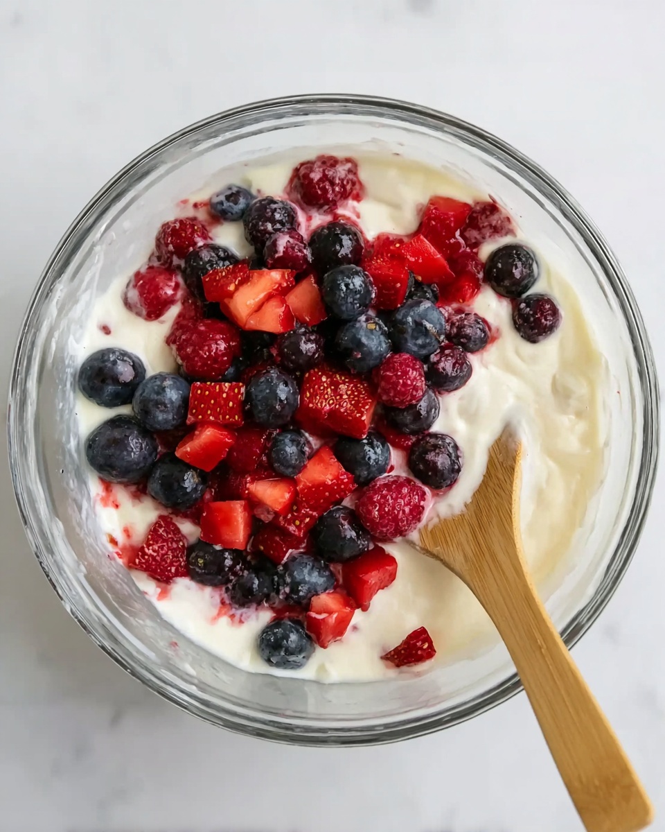 A clear glass bowl filled with a creamy white yogurt base, topped with three layers of fresh berries: whole dark blue blueberries, bright red chopped strawberries, and deep red raspberries scattered on top. There is a light wooden spoon resting in the mixture on the right side of the bowl, partially immersed in the yogurt and berries. The bowl sits on a white marbled surface, giving a clean and fresh look. Photo taken with an iphone --ar 4:5 --v 7