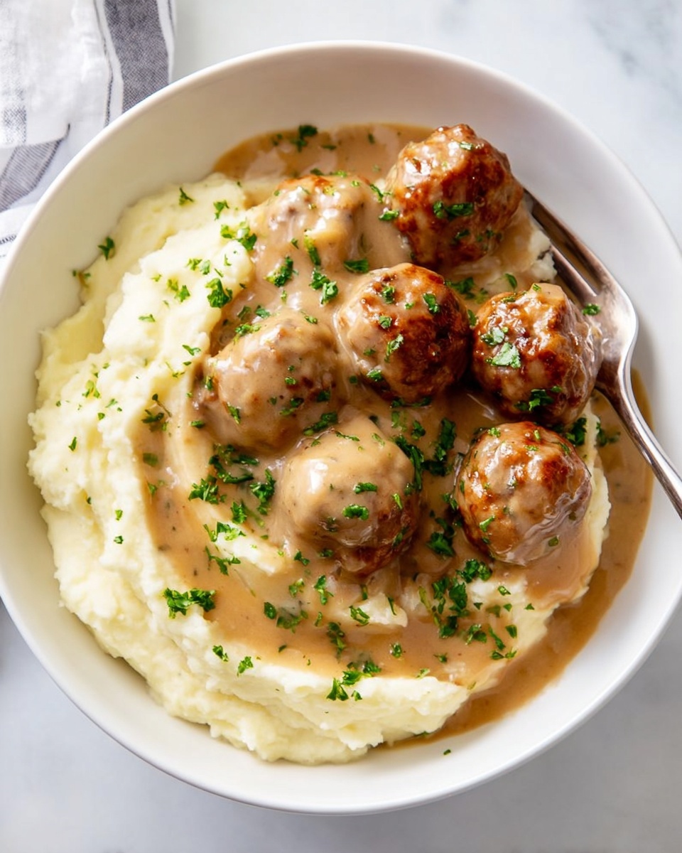 A white bowl sits on a white marbled surface, filled with a base layer of creamy mashed potatoes that are soft and smooth, occupying roughly half the bowl on the right side. On top and to the left, there are about seven round meatballs covered in a thick, light brown gravy that glistens slightly. The gravy also spills over and partially covers the mashed potatoes, adding a glossy texture. The dish is sprinkled with finely chopped green herbs, adding small pops of green color. A silver fork rests on the right edge of the bowl, partially on the mashed potatoes. photo taken with an iphone --ar 4:5 --v 7
