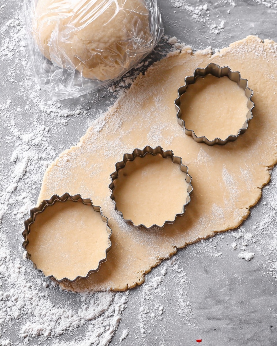 The image shows one layer of light beige dough rolled out thin on a white marbled surface dusted with white flour. Two scalloped round shapes are cut out from the dough with a metal round scalloped cutter placed on top of the dough. Above the dough, there is a wrapped ball of dough covered in clear plastic wrap, resting on the same white marbled surface. Photo taken with an iphone --ar 4:5 --v 7