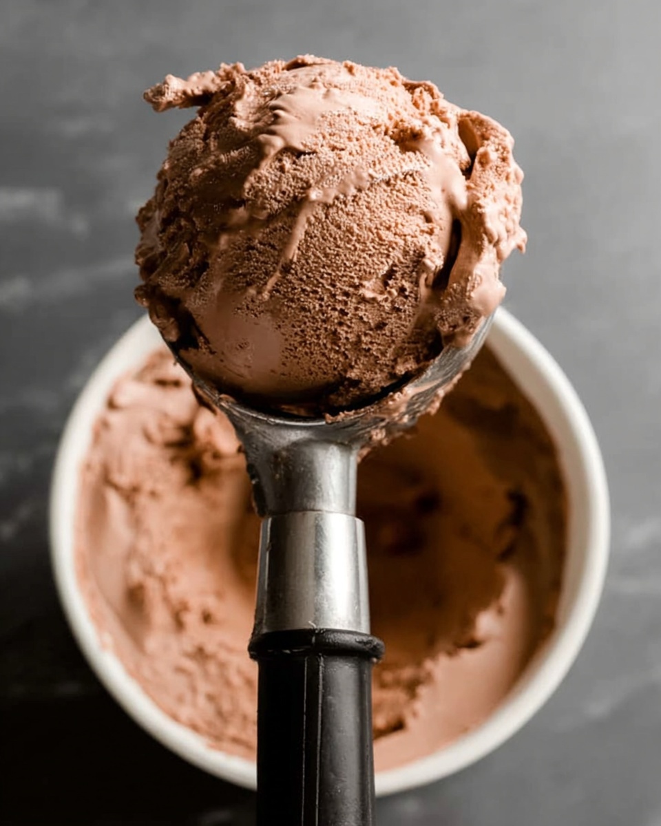 A close-up view of a rectangular white baking pan lined with parchment paper, filled with a thick layer of smooth, dark brown chocolate ice cream. A scoop is partially taken out, creating a rounded hollow with fine, creamy texture visible inside. The chocolate ice cream's surface shows slight ripples and a shiny, soft appearance. The background has a white marbled texture. Photo taken with an iphone --ar 4:5 --v 7