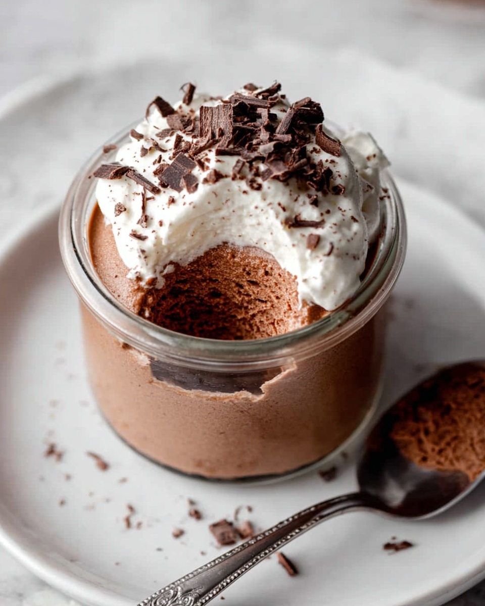 Three small clear glass jars are filled with smooth, creamy chocolate mousse that has a light brown color and silky texture. Each mousse jar is topped with a small dollop of white whipped cream that looks soft and fluffy. Dark chocolate shavings are sprinkled over the whipped cream and the mousse around it, adding a rich, textured contrast. The jars are placed on a large white plate with a soft white marbled surface beneath, and some loose chocolate shavings are scattered around the jars on the plate. A silver spoon is partially visible in the top right corner. photo taken with an iphone --ar 4:5 --v 7