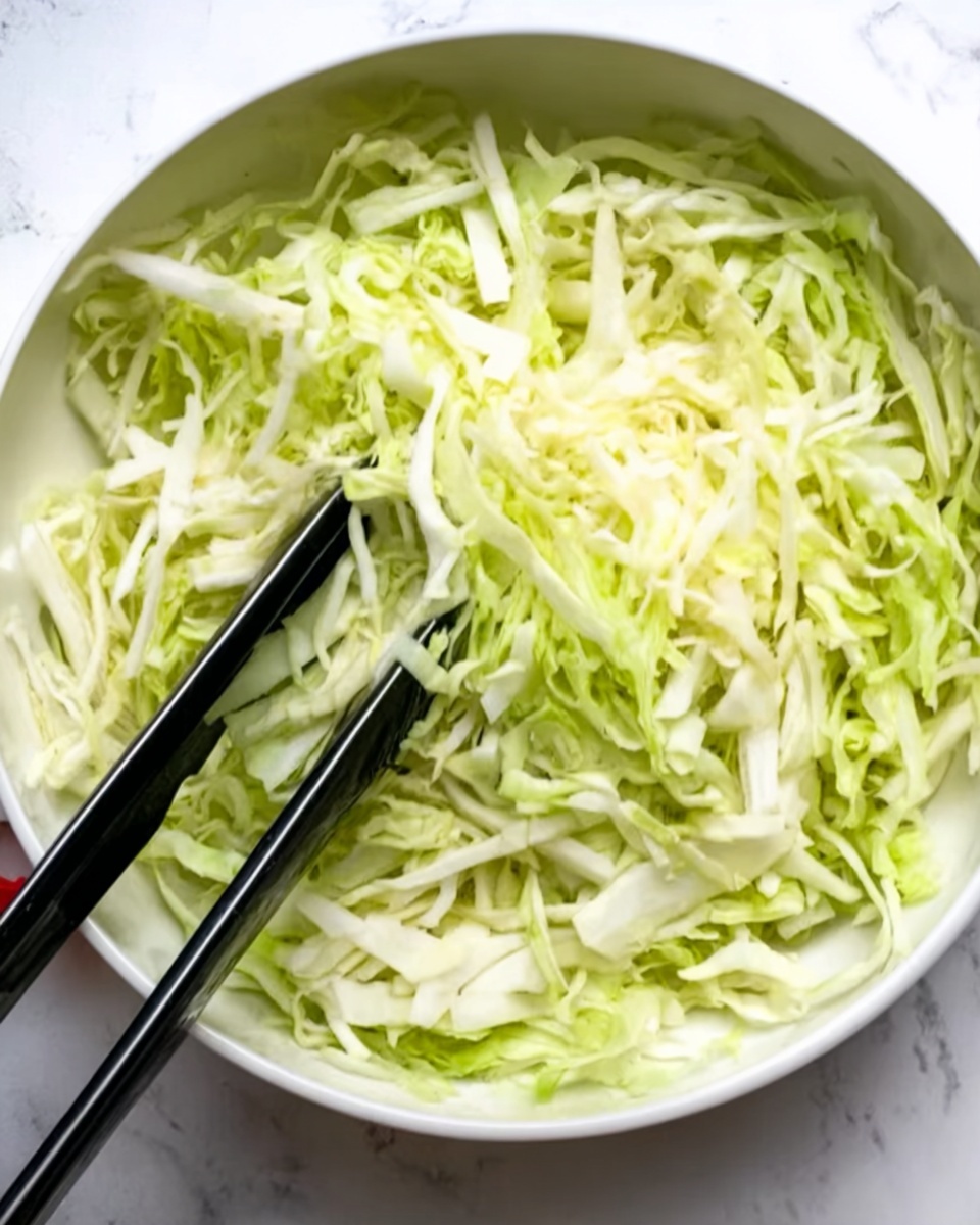 A white bowl filled with thinly sliced pale green and white cabbage, showing a mix of soft and crunchy textures. Two black tongs rest inside the bowl, one positioned diagonally across the bowl’s top and the other along the side, lightly touching the cabbage. The background is a white marbled surface. photo taken with an iphone --ar 4:5 --v 7