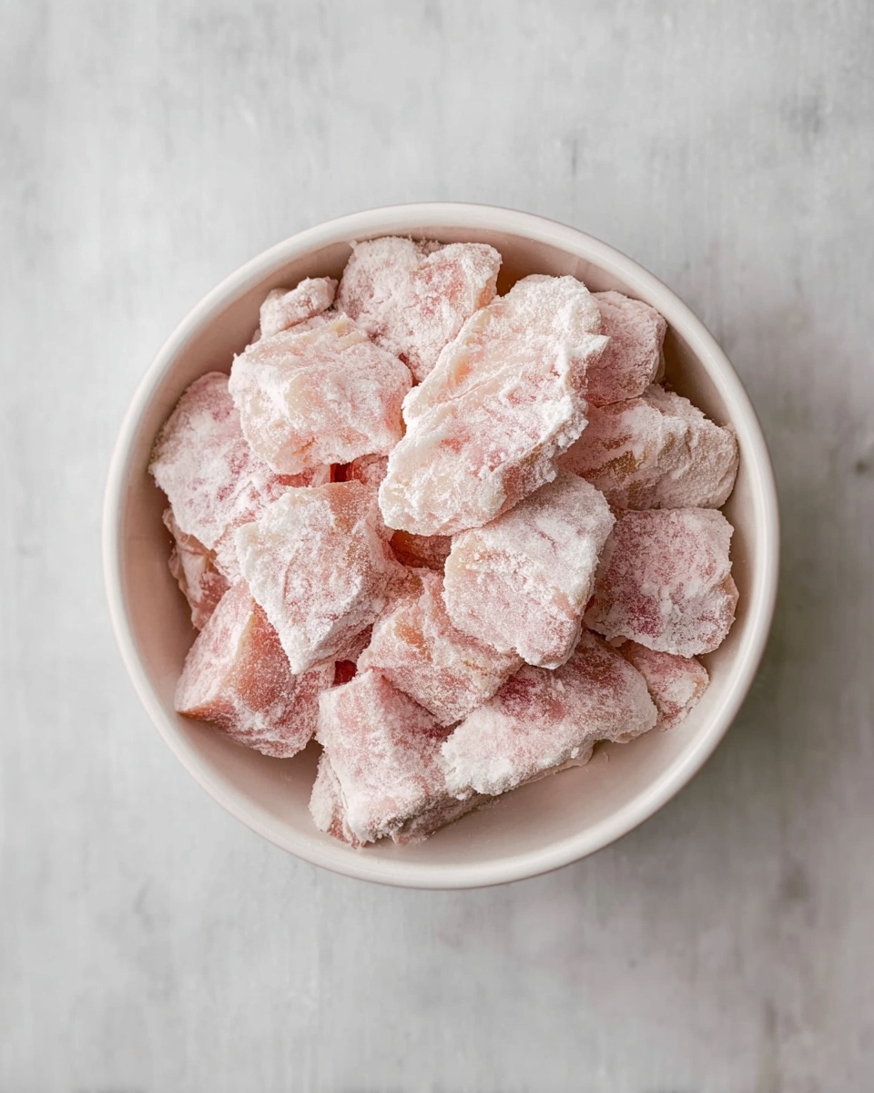 A white bowl filled with layers of frozen, pale pink chunks of meat dusted with a light white frost coating, each piece irregularly shaped and piled loosely. The bowl sits on a white marbled surface, viewed from above, showing the texture and icy coating clearly. photo taken with an iphone --ar 4:5 --v 7